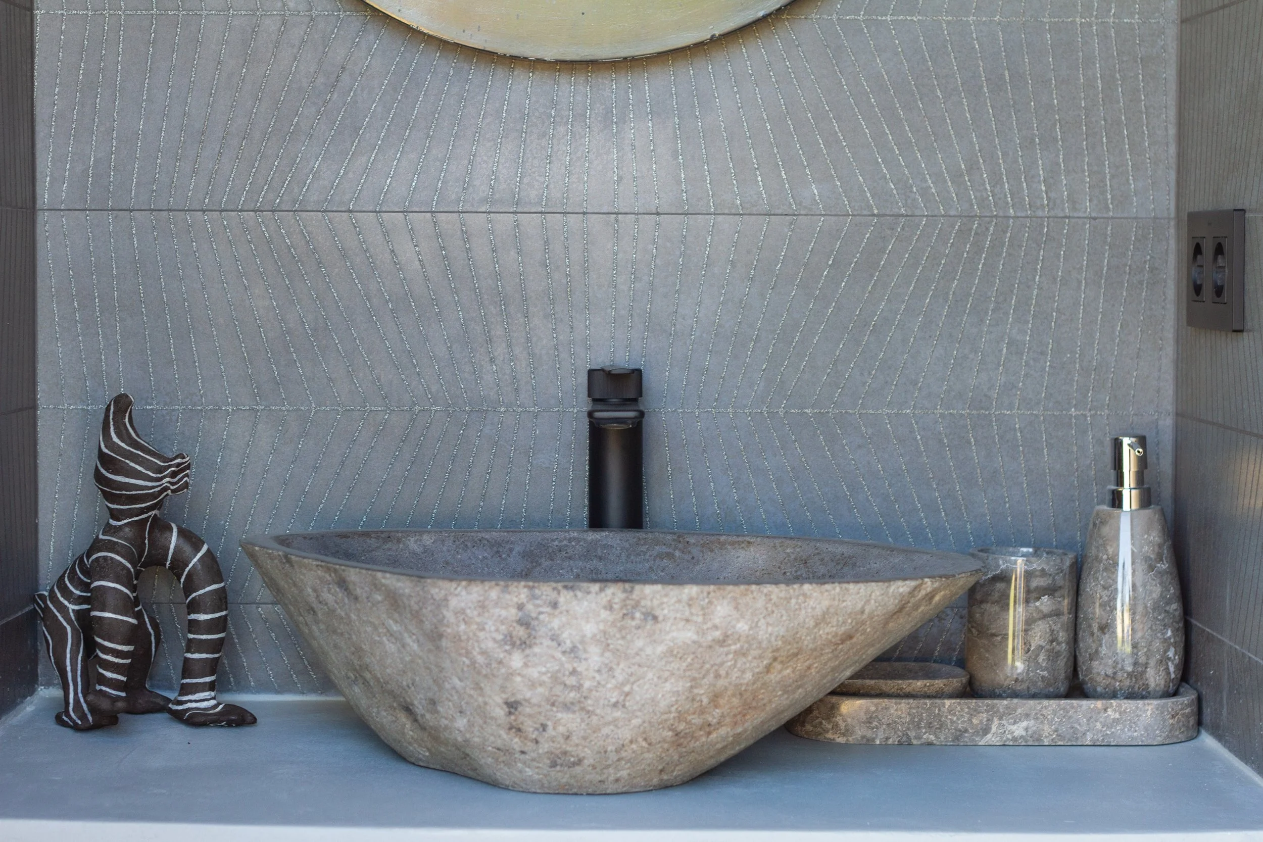 A decorative bathroom sink with a large, round stone basin, black faucet, and stone accessories including a soap dispenser and container. A striped decorative sculpture sits to the left of the sink, with a textured grey tile wall in the background.