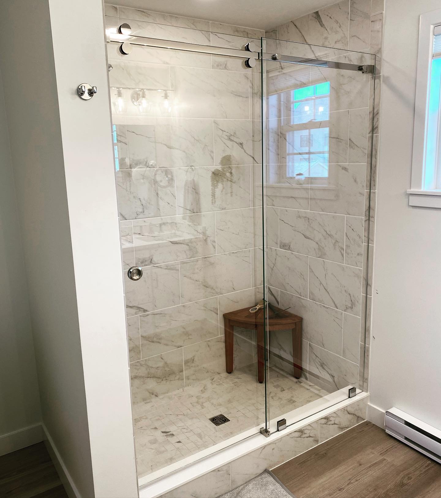 Low-iron glass Serenity barn-door-style sliding shower enclosure on a stainless top rail, with white marble-look tile and a teak shower stool, in a Scarborough Downs, Maine bathroom
