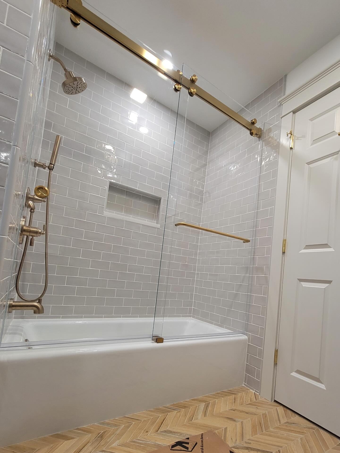 Low-iron frameless Cambridge bypass sliding glass door with brushed bronze rail and towel bar over a tub with gray subway tile and a niche in a Kennebunk, Maine bathroom