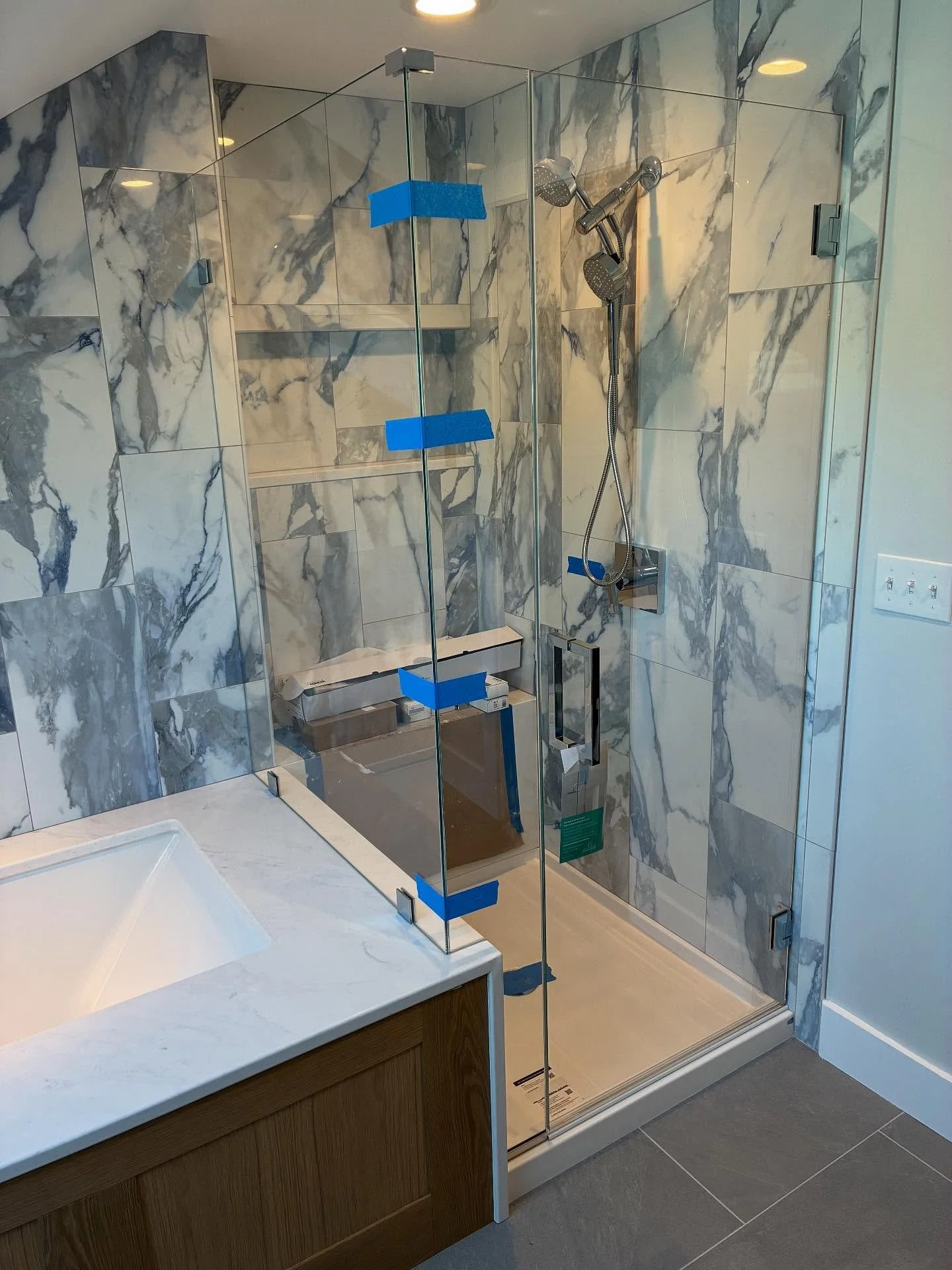 Frameless low-iron glass shower enclosure with a hinged door and return panel, glass shelves and a bench, against gray and white marble-look tile beside a vanity in a Cumberland, Maine bathroom