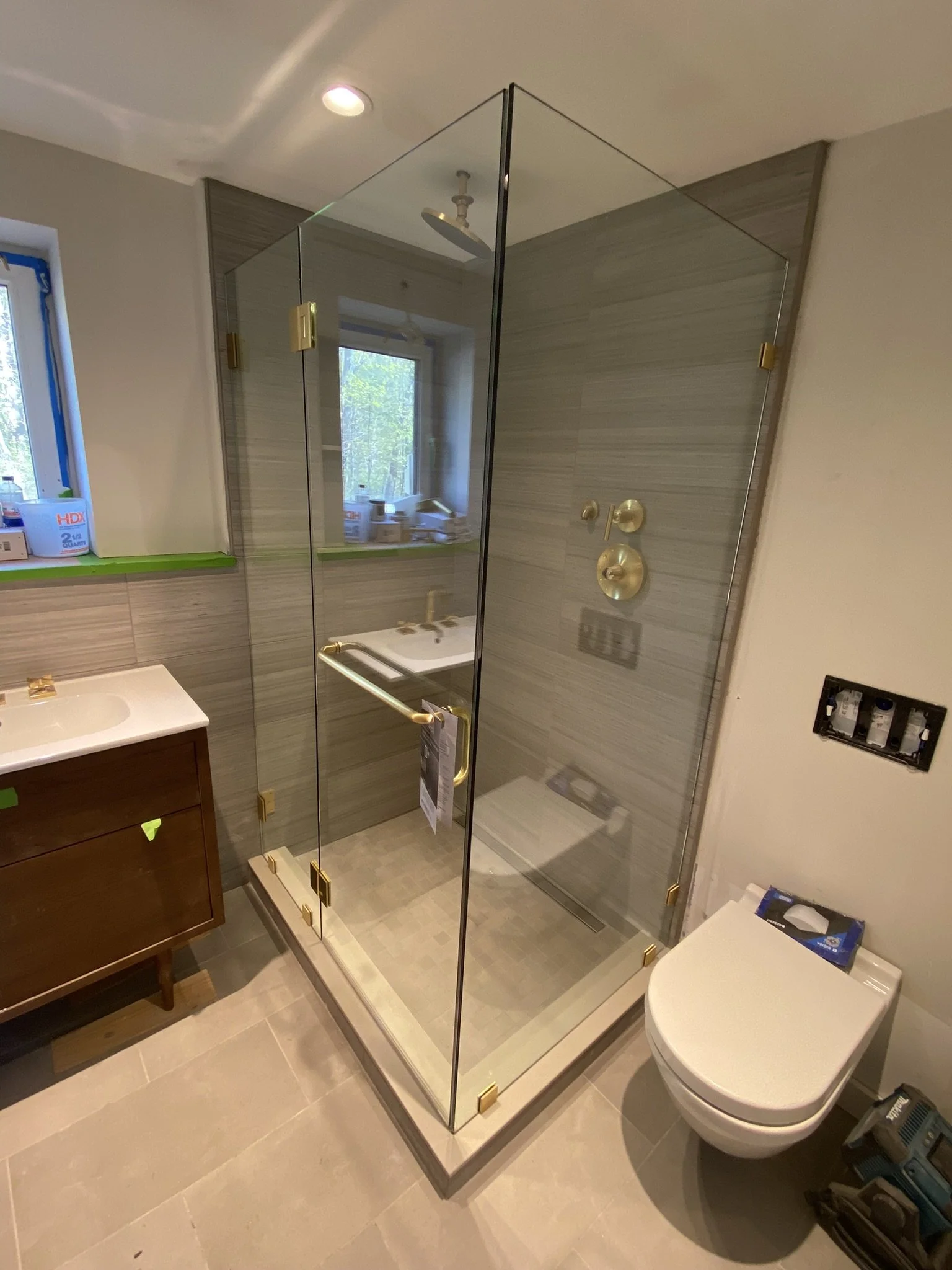 A modern bathroom with a glass shower enclosure, a window, a wooden vanity with a white sink, and a wall-mounted toilet. The shower has gold fixtures and hinges, and the bathroom has tiled walls and floors.