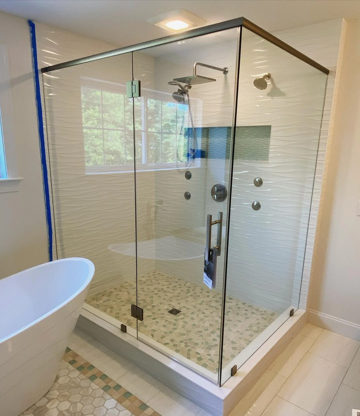 Large glass corner shower with a header bar, white wave tile and pebble floor beside a freestanding tub in Saco, Maine