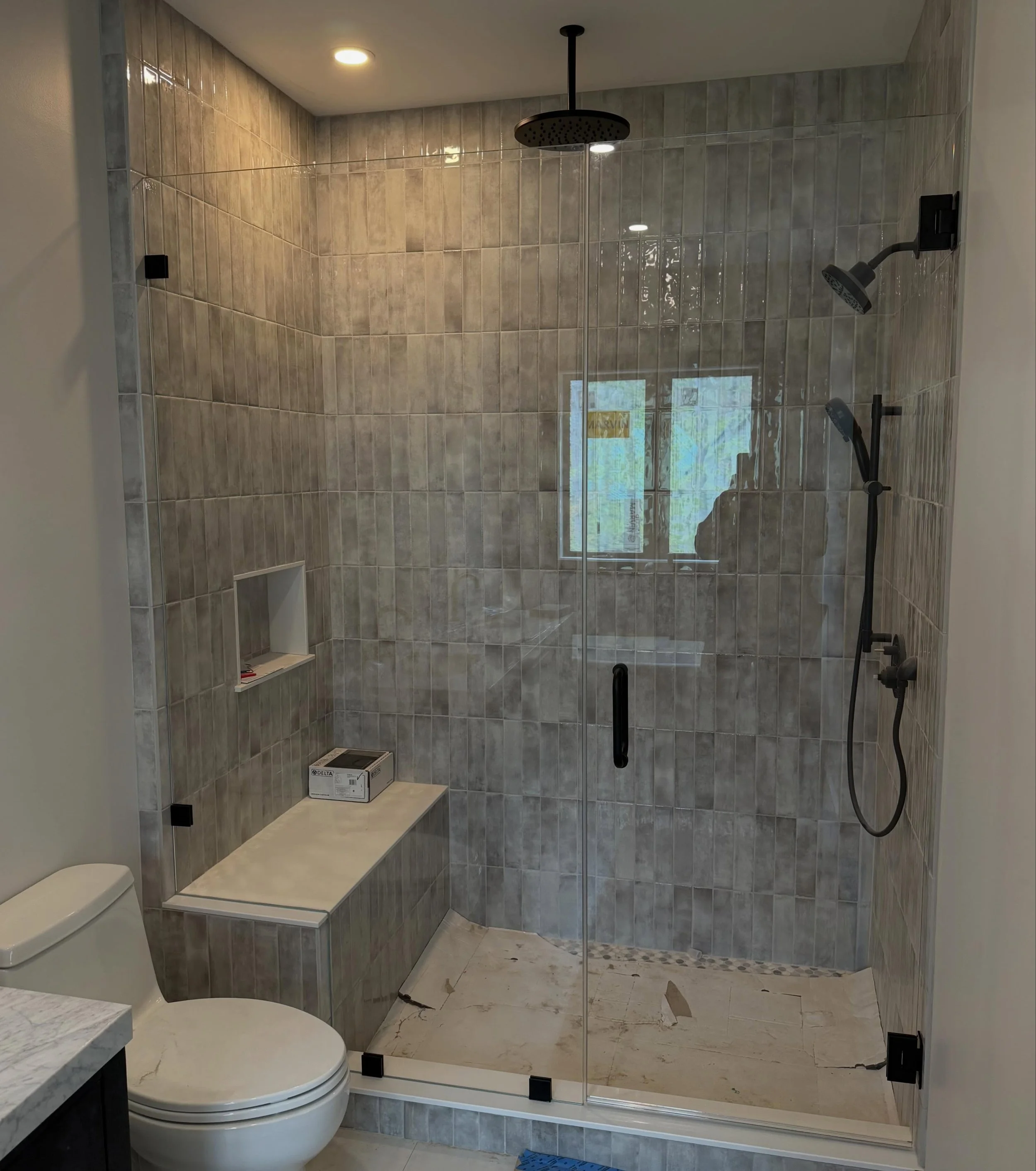Spacious frameless low-iron Starphire glass shower with a hinged door and panel, matte black hardware, a tiled bench, a window and a ceiling rain head, in a Naples, Maine bathroom