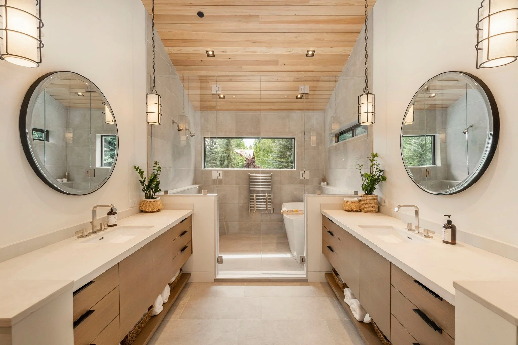 Modern double vanity bathroom with large round mirrors, potted plants, pendant lights, and a glass-enclosed shower area at the back with a window showing greenery outside.