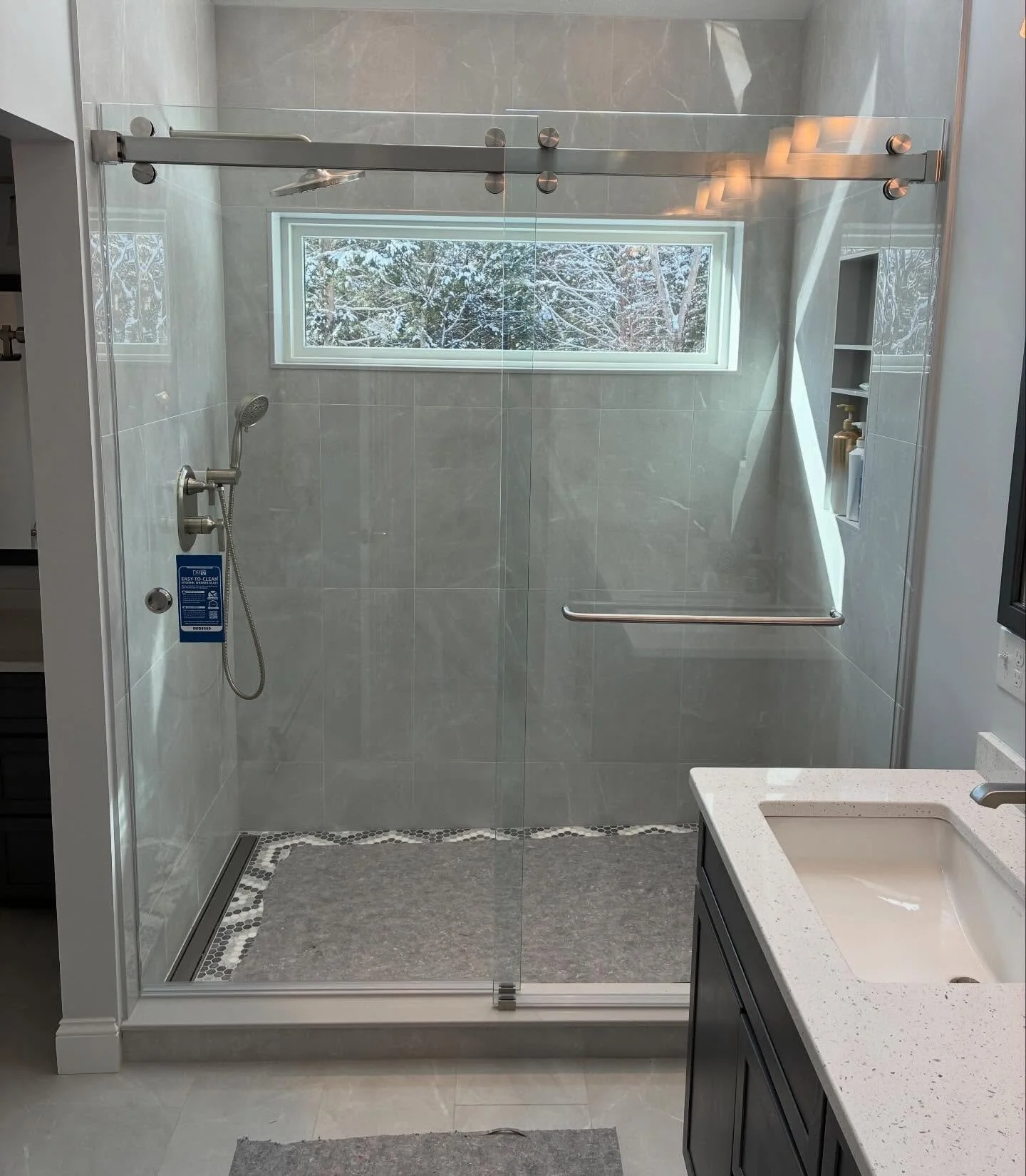 CRL Cambridge frameless sliding glass shower door on a brushed stainless top rail, with a transom window, gray tile walls and a pebble floor, in a Scarborough, Maine bathroom