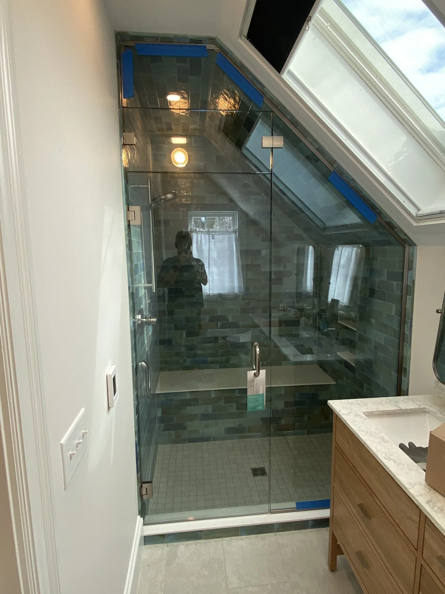 A bathroom with a glass shower enclosure under a sloped ceiling with a skylight, tiled blue-green walls, a white countertop with a wooden cabinet, and a wall-mounted mirror.