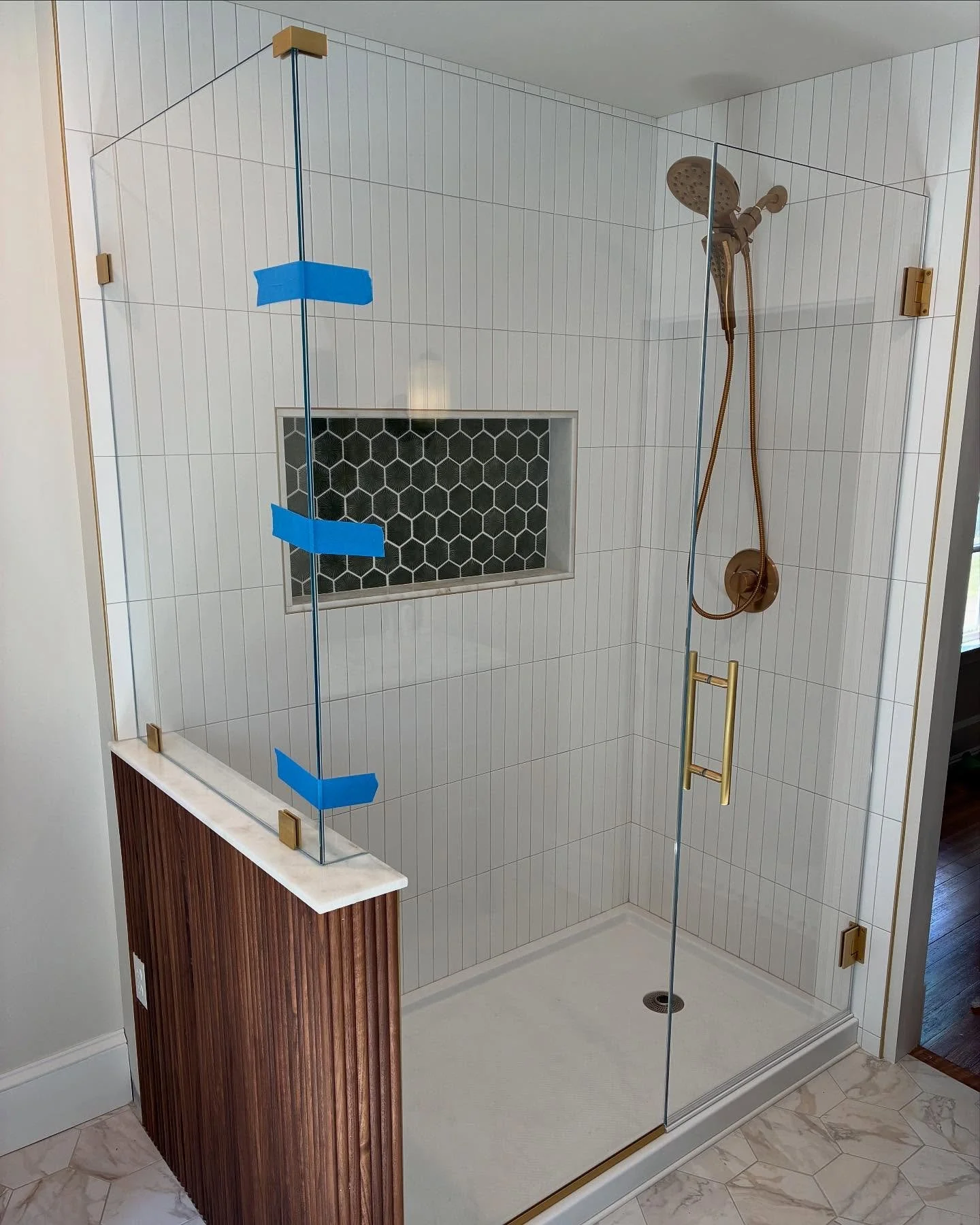 Frameless low-iron glass shower enclosure with a hinged door and a panel on a wood-paneled knee wall, gold-tone hardware and fixtures, white tile and a black hexagon niche in a South Portland, Maine bathroom
