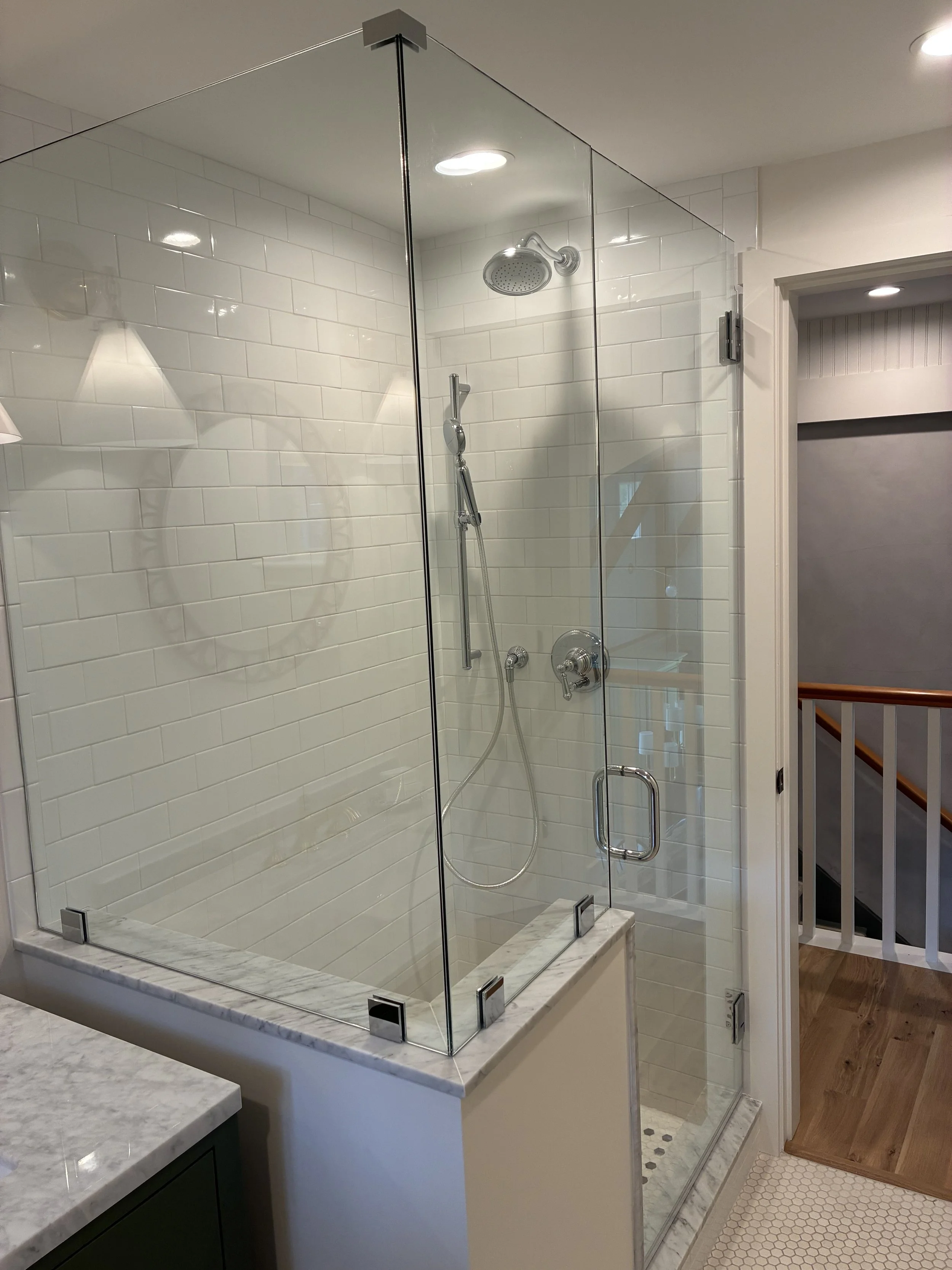 A glass-enclosed shower with white subway tiles, a rainfall showerhead, a handheld showerhead, and a soap dish, next to a hallway with wooden flooring and a staircase.