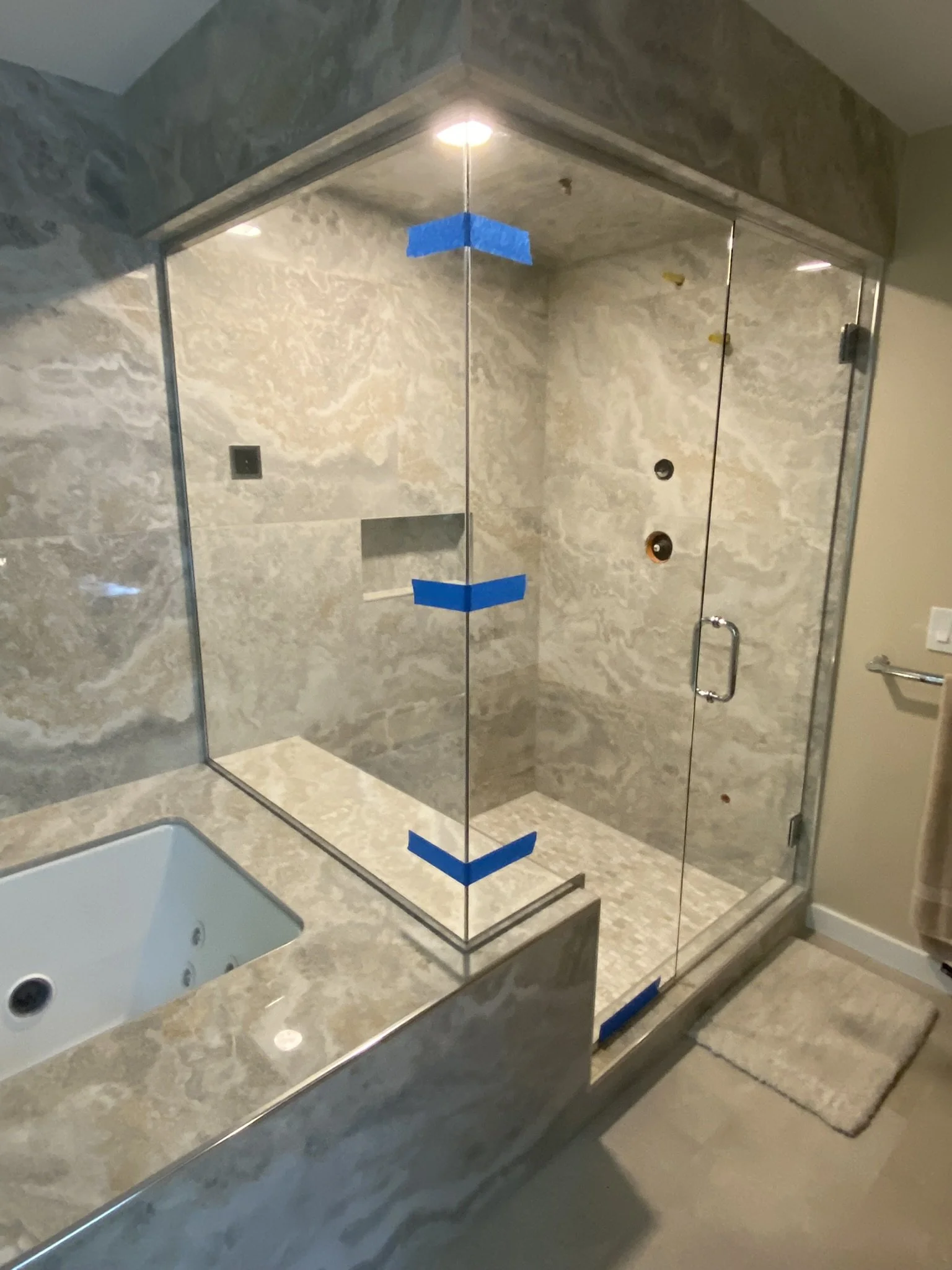 A glass-enclosed shower with marble walls and a marble bathtub nearby in a modern bathroom, with a beige bath mat on the floor.