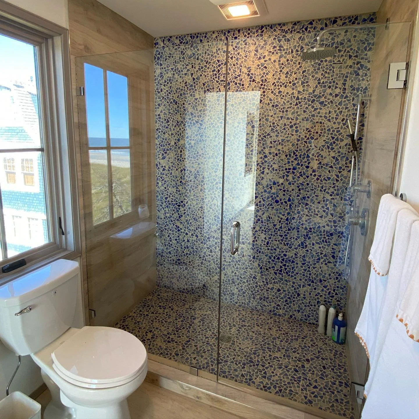 Frameless glass shower door and inline panel with polished chrome hardware and blue and tan sea-glass mosaic tile in a Biddeford Pool, Maine bathroom with an ocean view