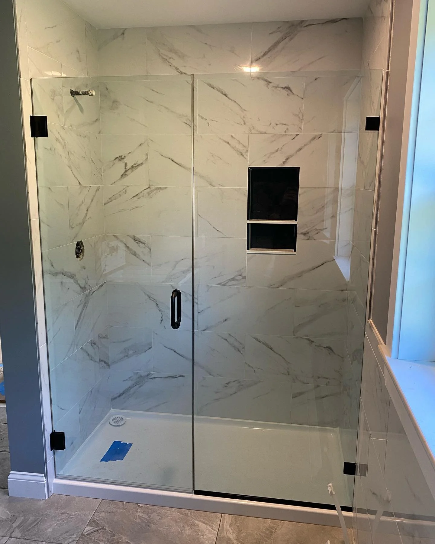 Frameless glass shower door and inline panel with matte black hardware on a white shower base, with white marble-look tile walls and a black niche, in a Standish, Maine bathroom