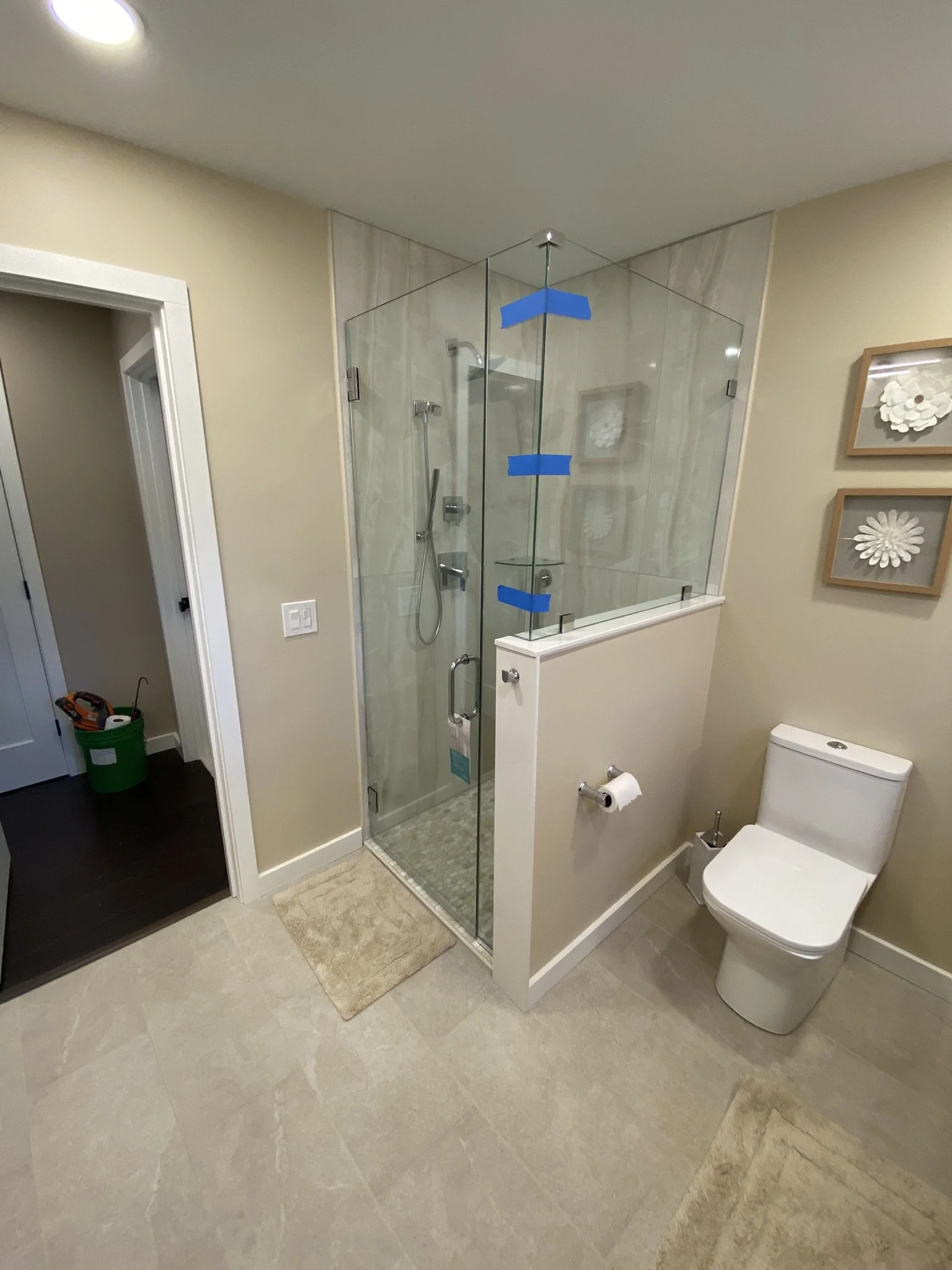 A bathroom with a glass shower enclosure, a white toilet, beige walls, and wall art with floral designs.
