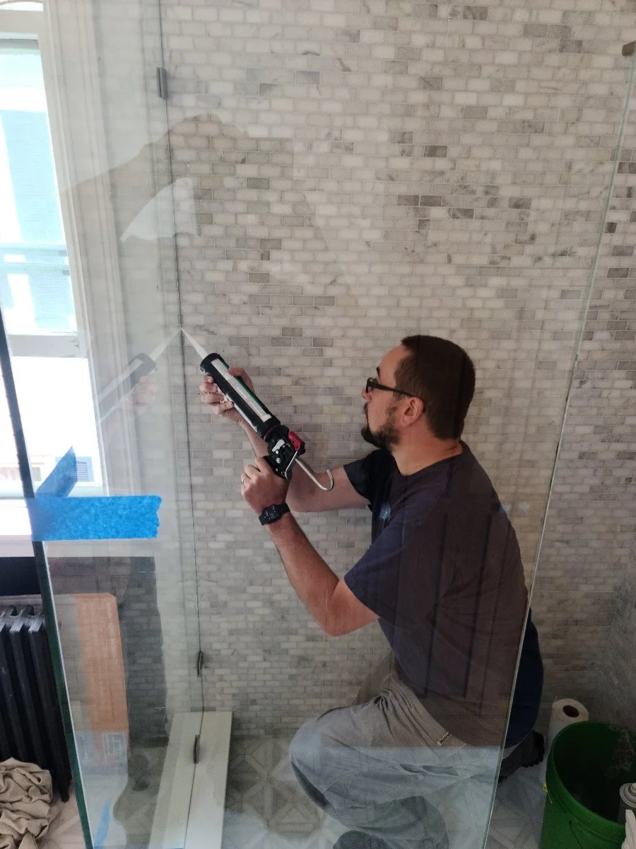 A man with glasses and a beard using a caulking gun inside a glass shower stall, with a stained glass window nearby and a green bucket on the floor.