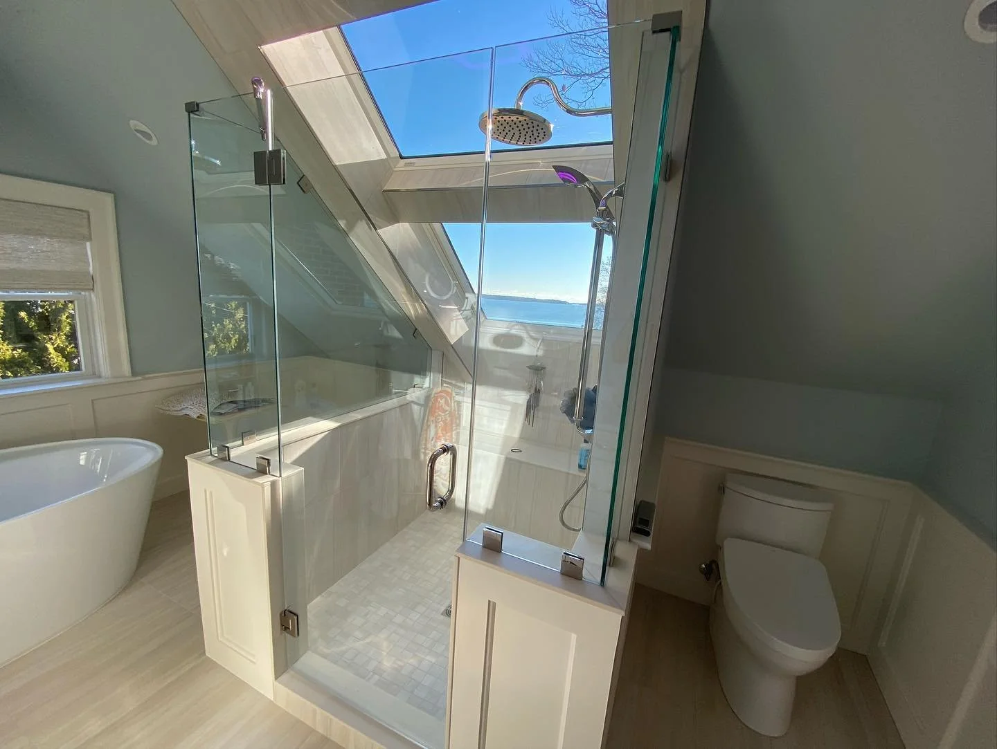 Frameless glass shower enclosure on white knee walls under a sloped ceiling and skylight with an ocean view, beside a freestanding tub in a Falmouth Foreside, Maine bathroom