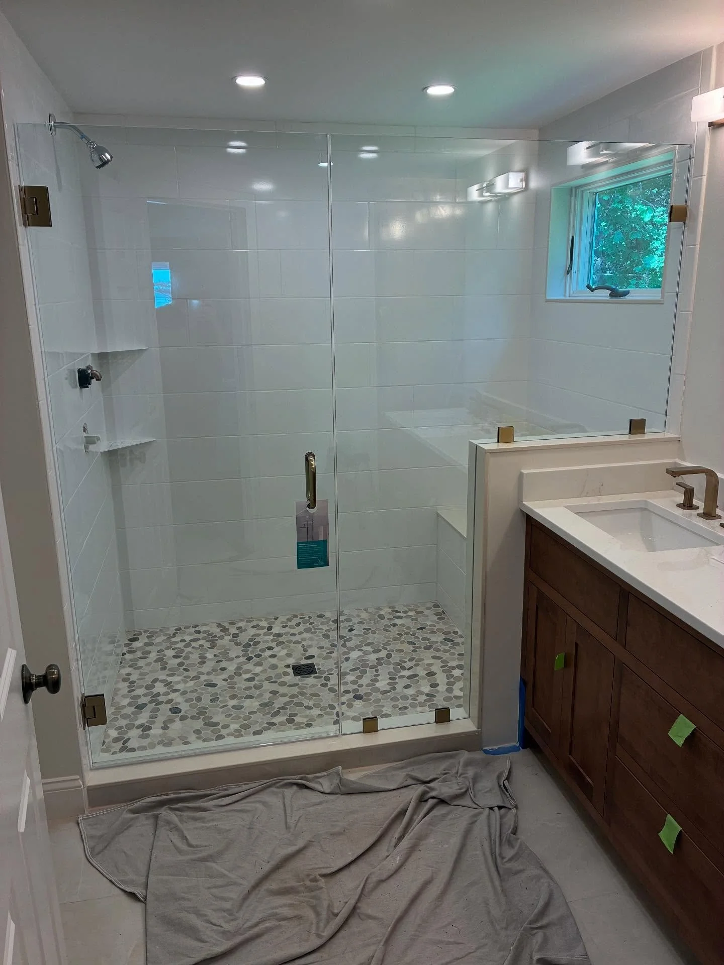 Frameless glass shower door, fixed panel and short return on a tiled knee wall with brushed bronze hardware, white tile, pebble floor and bench in a Biddeford Pool, Maine home
