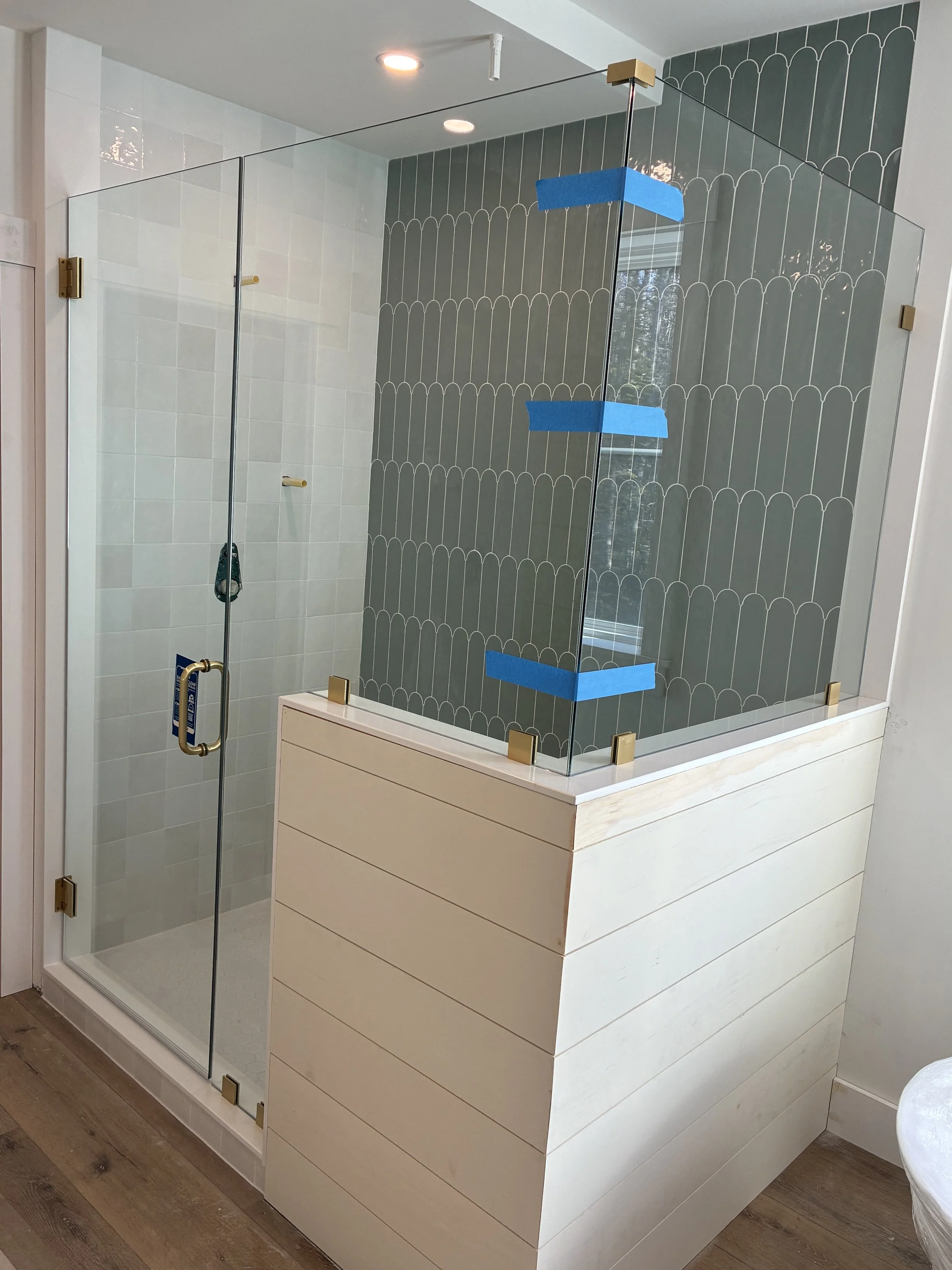 A modern shower enclosure with glass walls, a wooden base, and decorative tile wall. The glass walls are held with gold-colored brackets, and blue painter's tape is on the glass. Inside the shower, there's a white tiled wall and a small hand towel ha