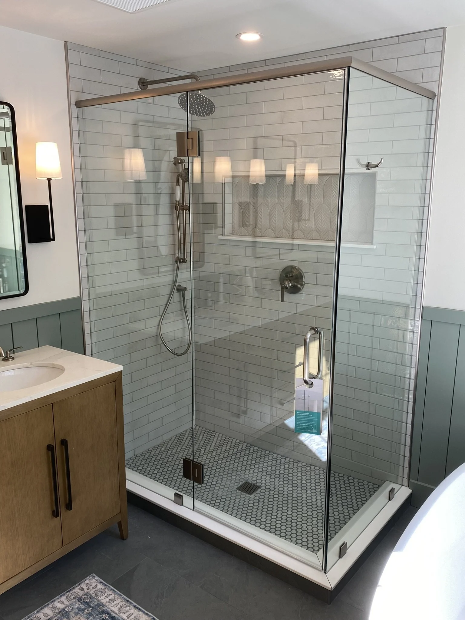 A modern bathroom with a glass shower enclosure, white subway tile walls, a rainfall showerhead, and a small window. To the left, there is a wooden vanity with a white countertop and a round sink, and a mirror and wall-mounted light fixture above.