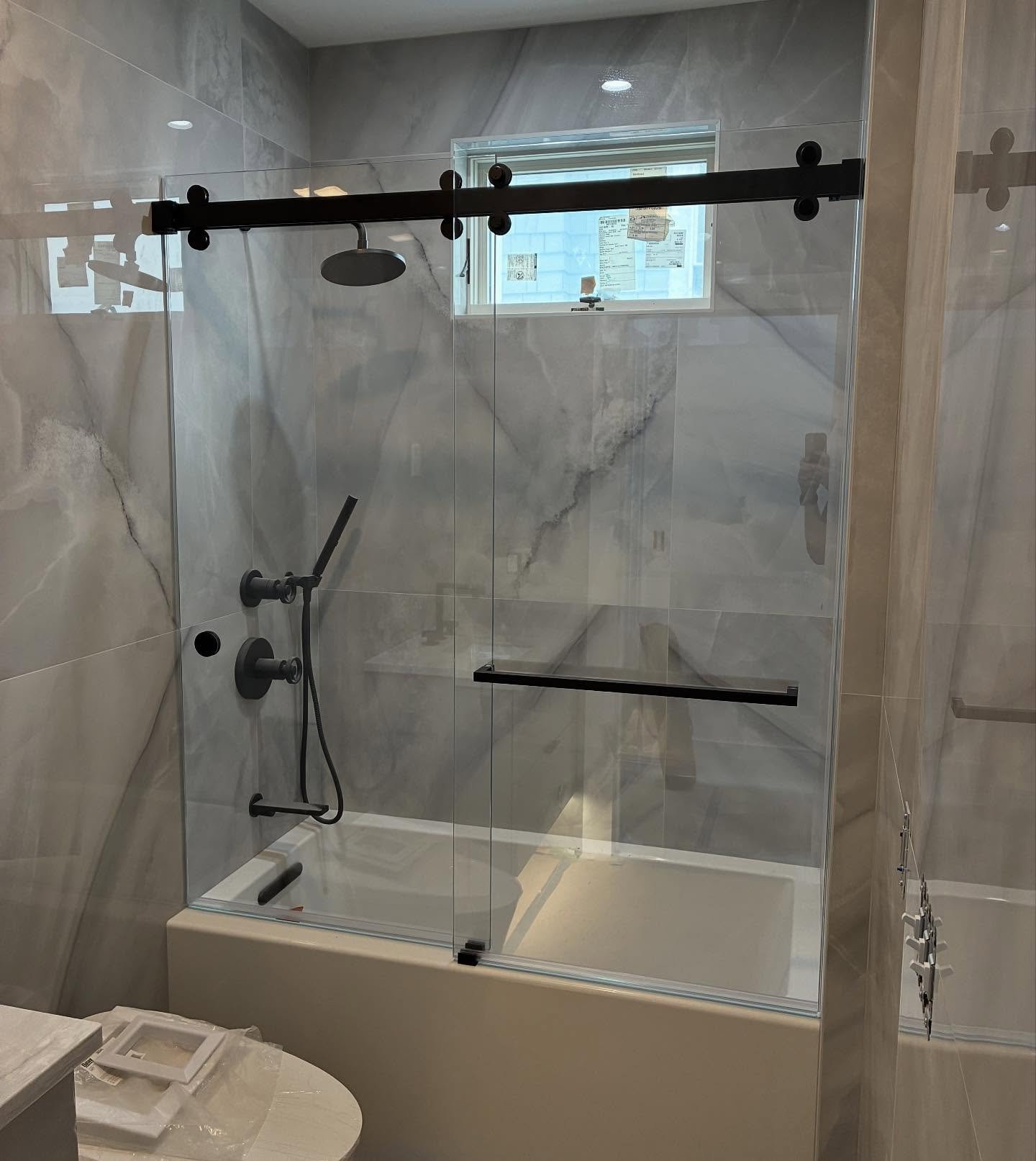 Low-iron CRL Cambridge bypass sliding glass door with a matte black top rail and towel bar over a tub, with gray marble-look tile and a window, in a Wells Beach, Maine bathroom