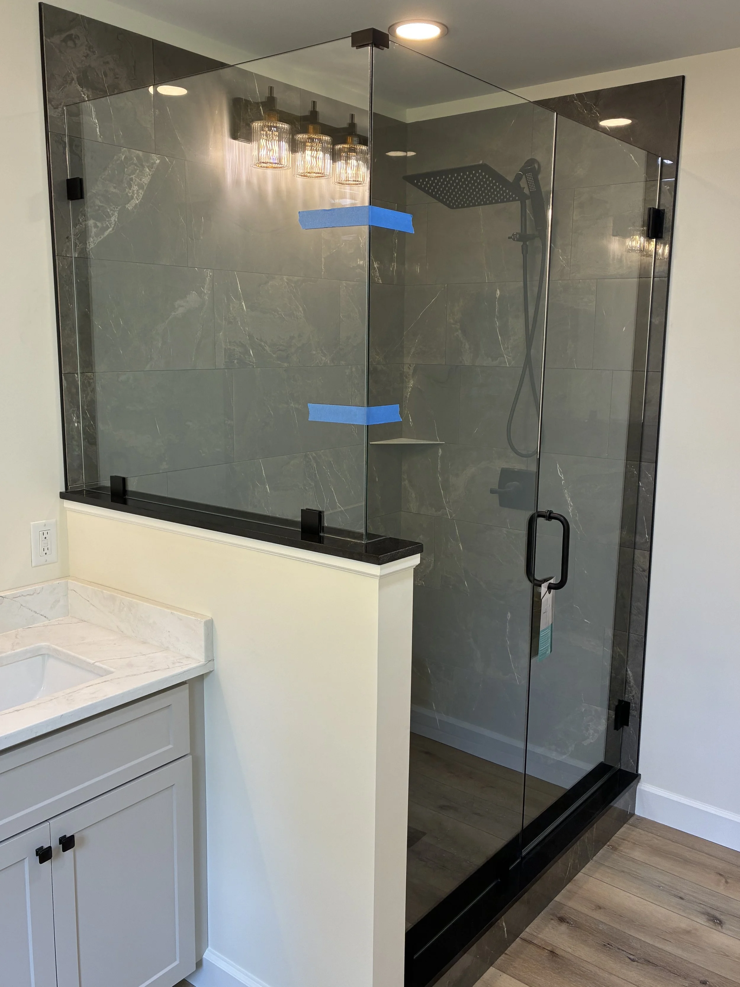 Modern glass shower enclosure with black fixtures, including a rainfall showerhead, in a bathroom with white walls and wood flooring.