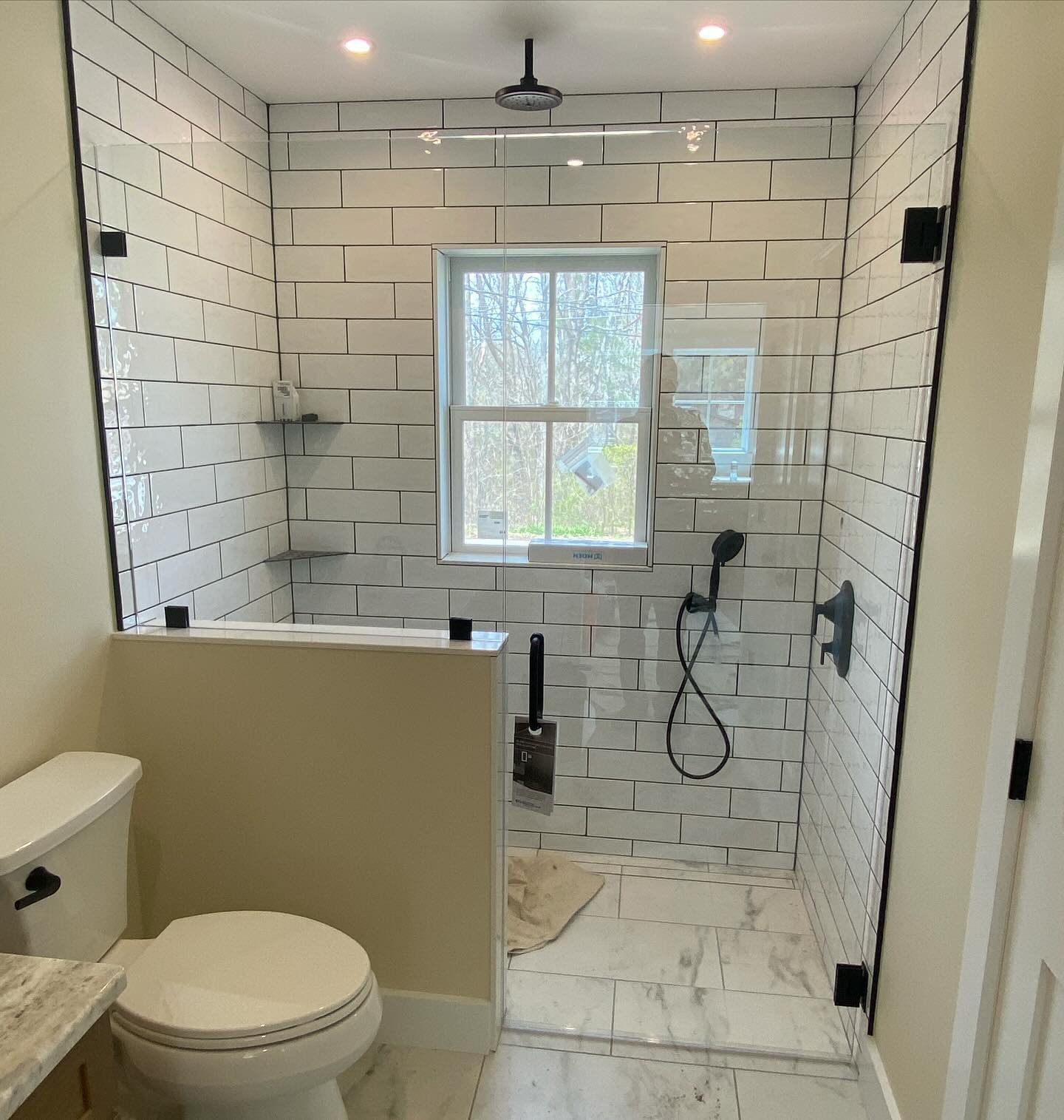 Frameless glass panels with matte black clips, one set on a half wall, on a curbless white subway tile shower with a window and black fixtures in a South Portland, Maine bathroom