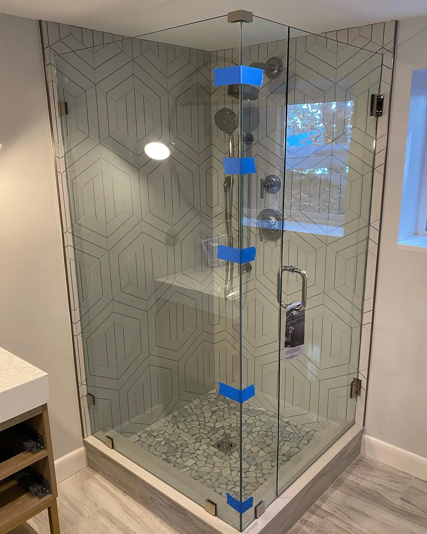 Frameless glass corner shower enclosure with chrome hardware, geometric white tile, a window and a pebble floor, in a Naples, Maine bathroom
