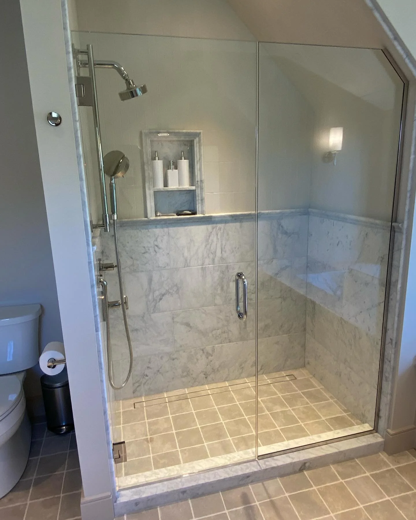 Frameless glass shower door and inline panel cut to a sloped ceiling over a marble tile shower with a linear drain in a Cape Elizabeth, Maine bathroom