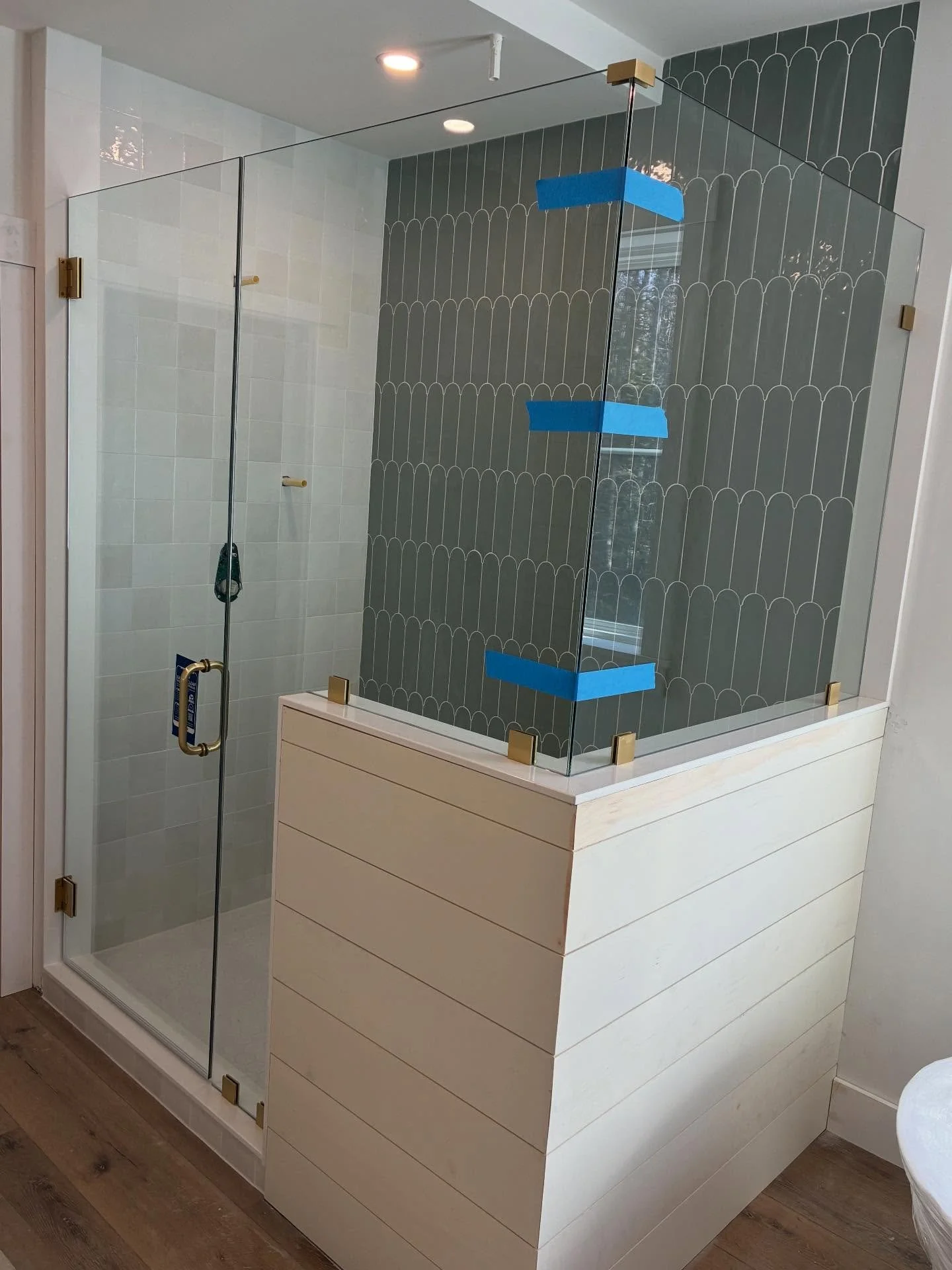 Frameless glass corner shower enclosure with a hinged door and glass panels set on a white shiplap knee wall, gold-tone hardware, and green fan-pattern accent tile in a Kennebunk, Maine bathroom