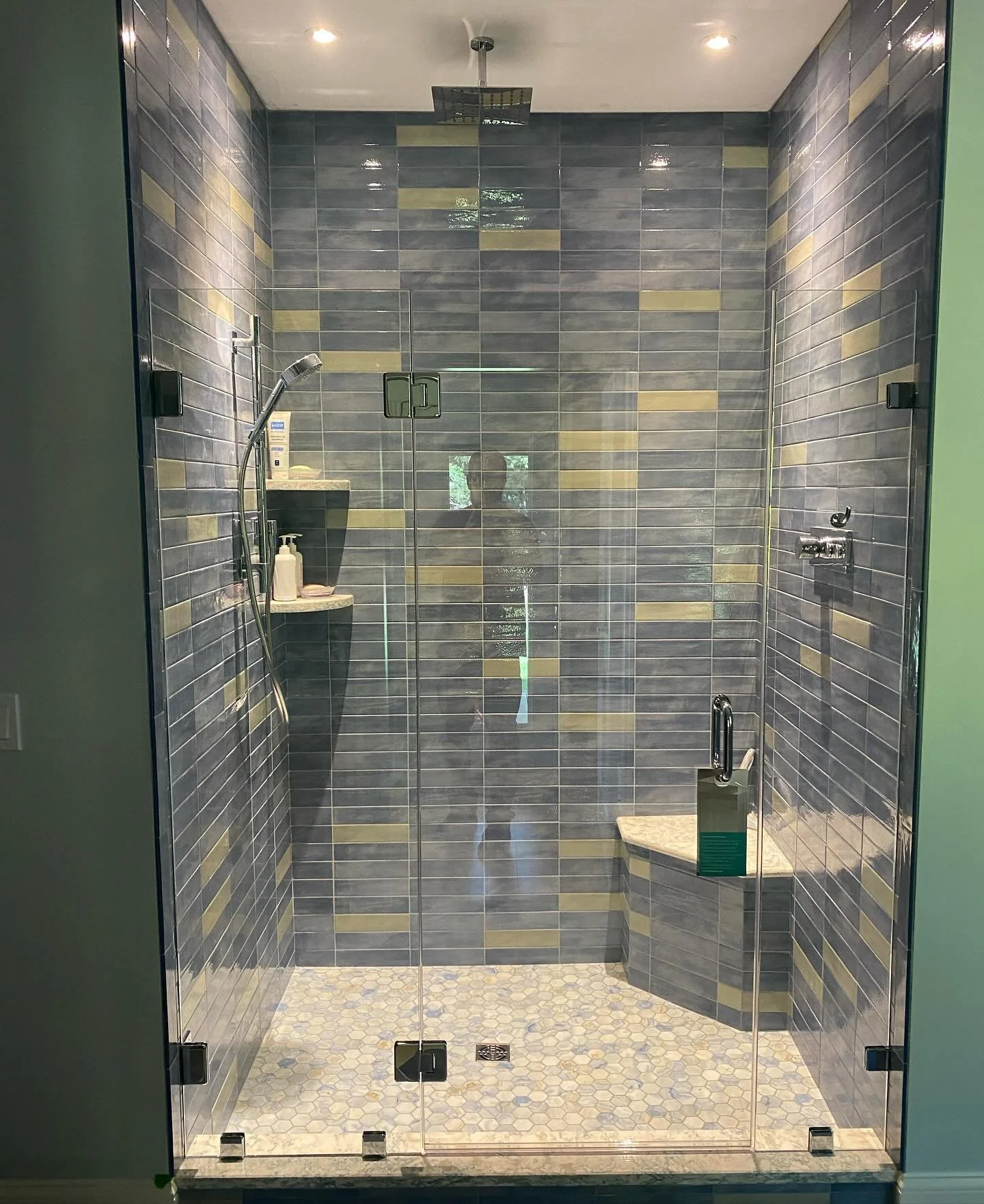Frameless glass shower door and inline panels across a blue and gold stacked tile shower with a corner bench, niches and pebble floor in a Cumberland Center, Maine bathroom