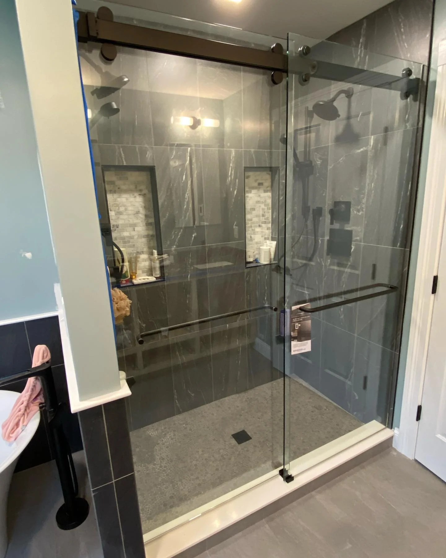 Frameless Cambridge sliding glass shower enclosure on a dark top rail with a towel bar, in front of a gray tile shower with twin niches in a Cumberland, Maine bathroom
