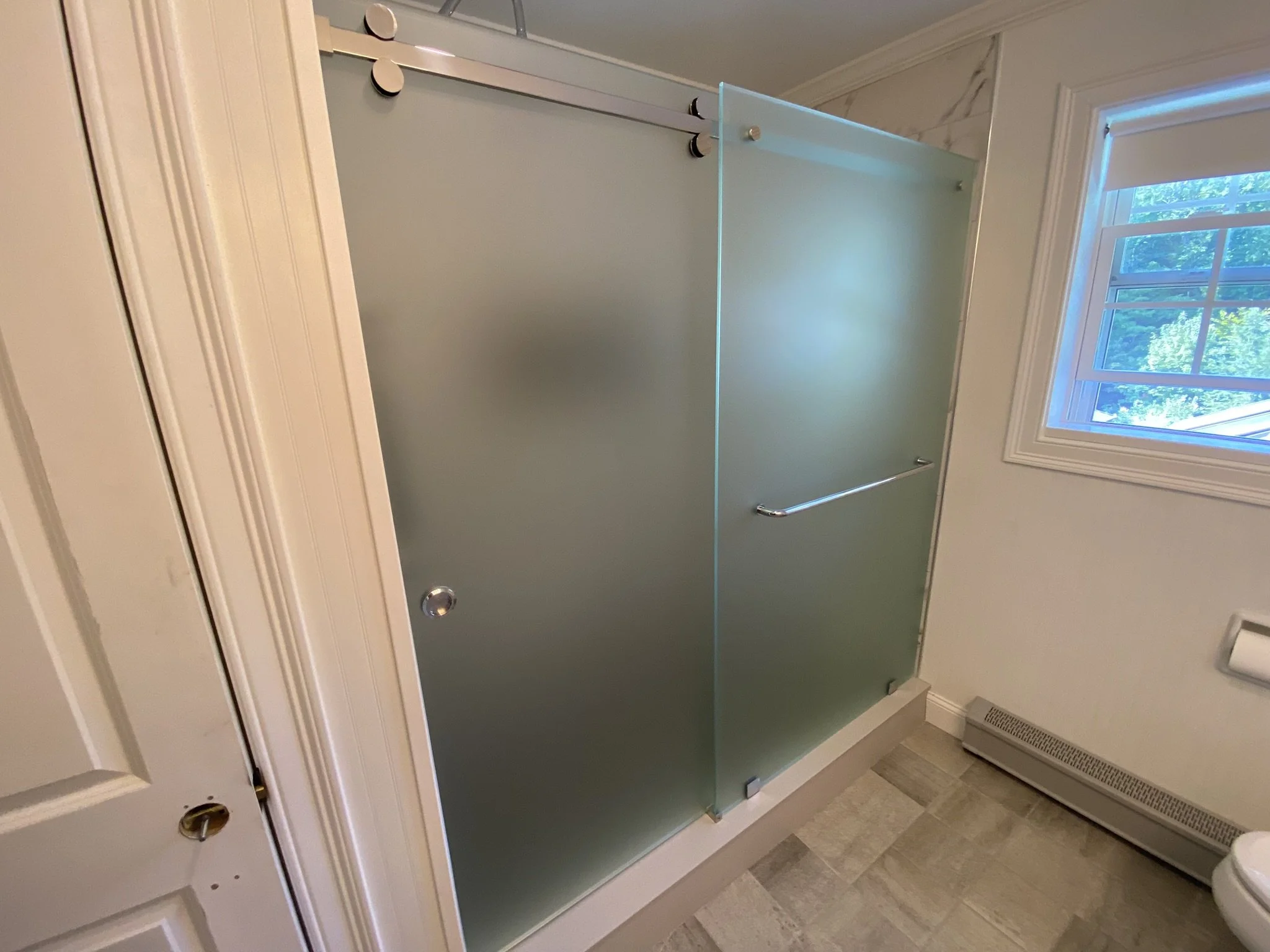 Frosted glass shower door with sliding mechanism in a bathroom, next to a window and a wall heater.