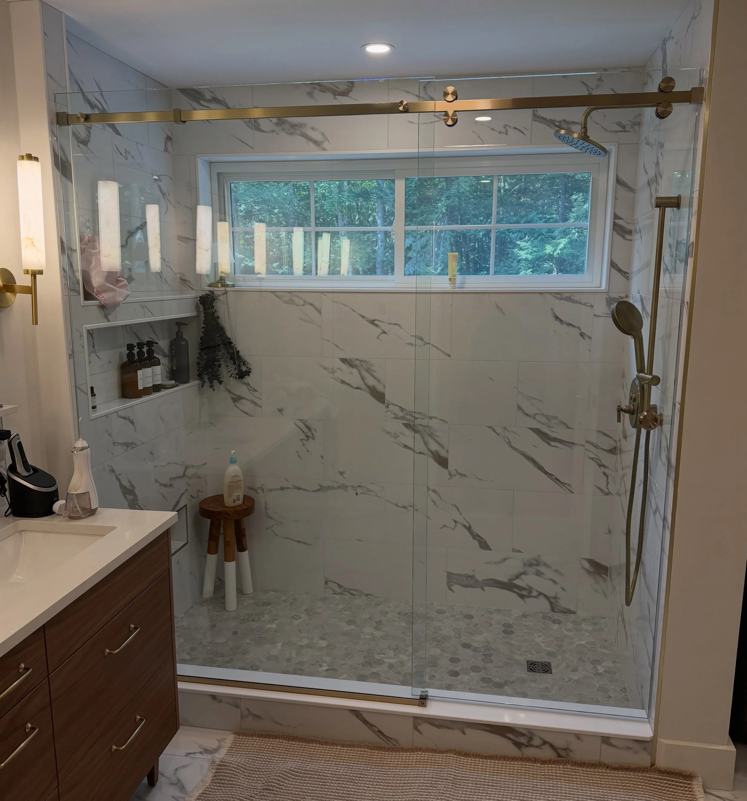 Frameless sliding glass shower door on a gold-tone top rail across a white marble-look tile shower with a window, niche and teak stool in a Falmouth, Maine bathroom