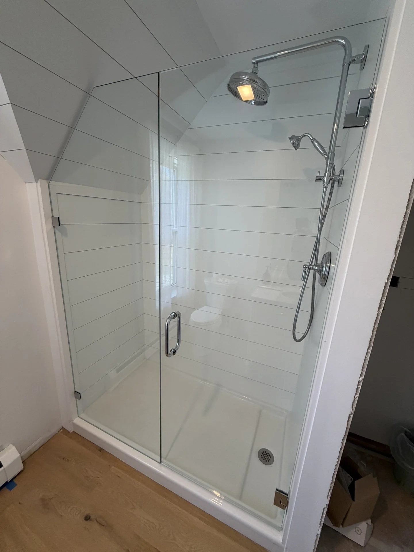 Glass shower door and fixed panel cut to a sloped ceiling over a white shower base, with white shiplap walls and a chrome shower system, in a Waterboro, Maine bathroom