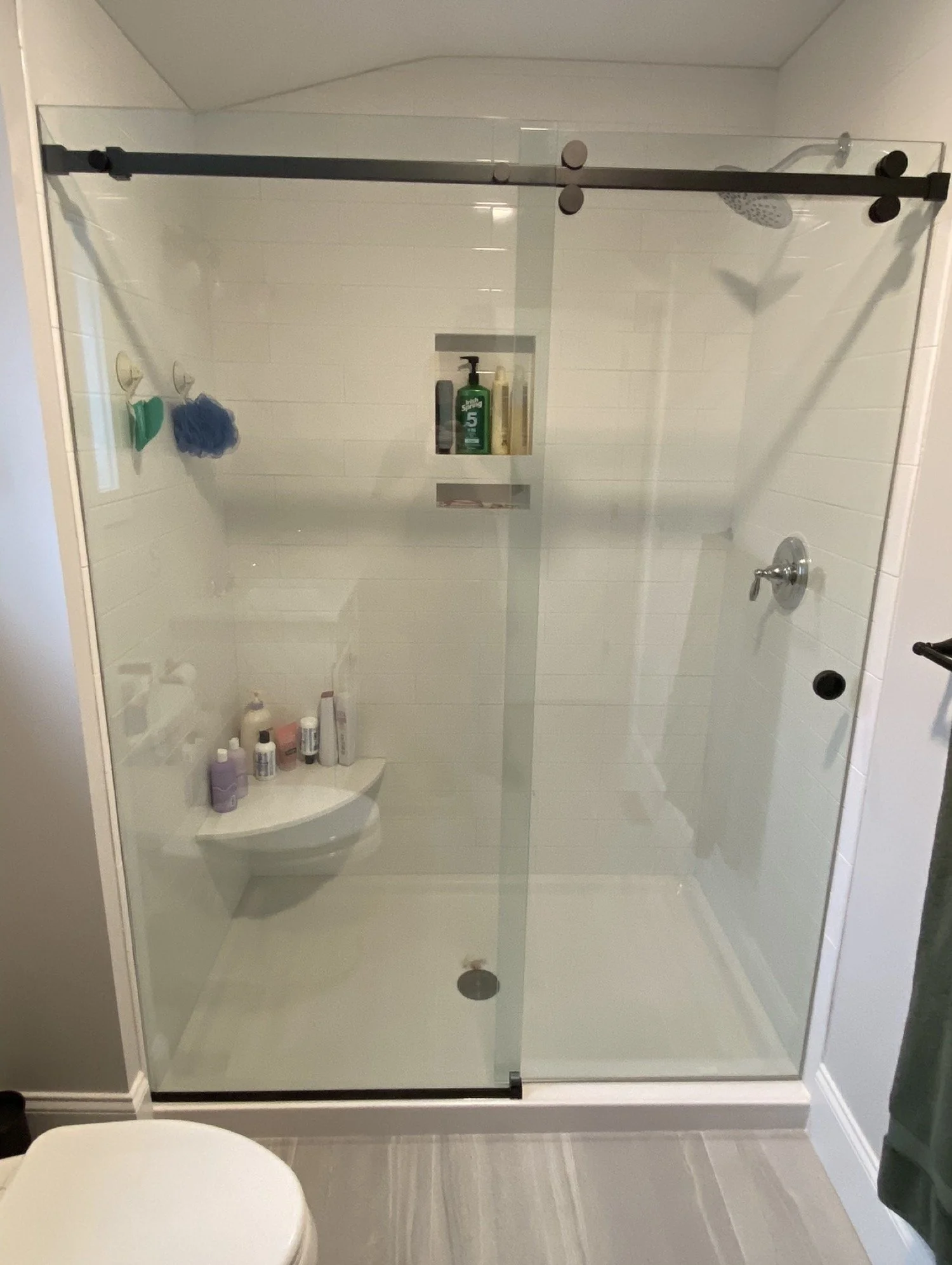 Frameless sliding glass shower door on a matte black roller rail in a white walk-in shower with a corner shelf, in an Ogunquit and York area Maine bathroom