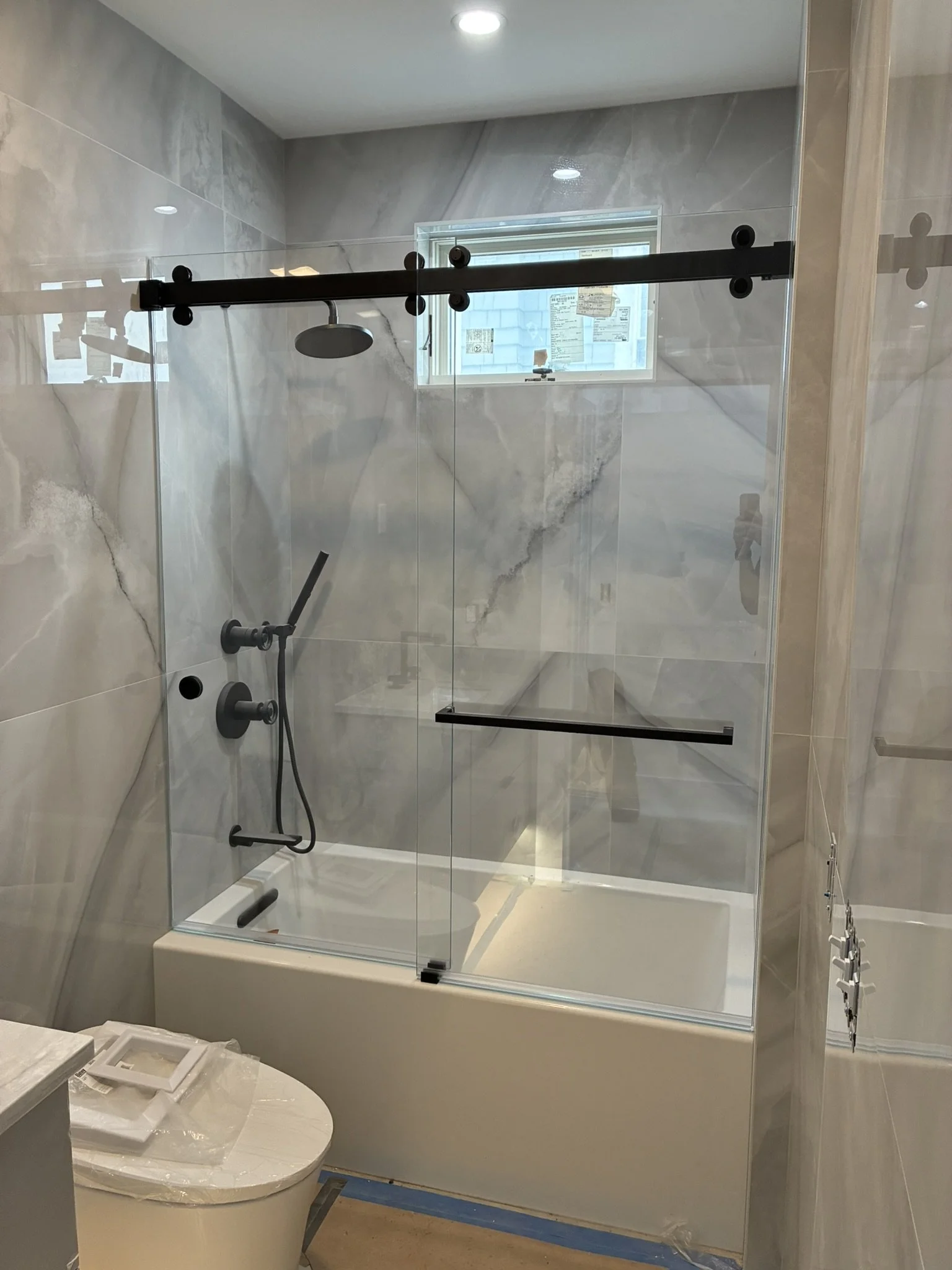 Modern bathroom with a glass shower enclosure, marble walls, a small window, and a toilet in the foreground.