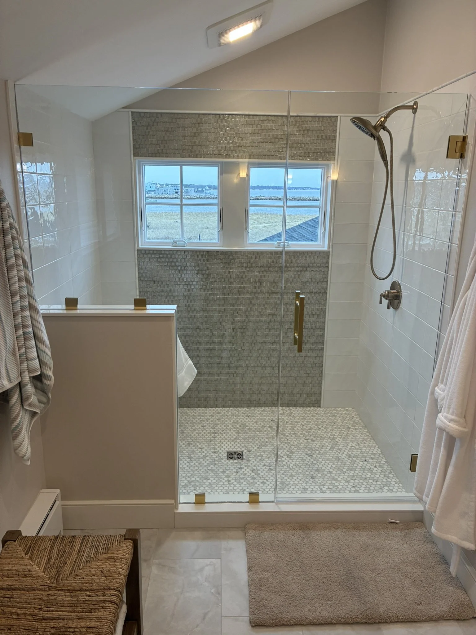 Modern walk-in shower with glass doors, tile wall and floor, and a window providing a view of an outdoor landscape with water and sky.