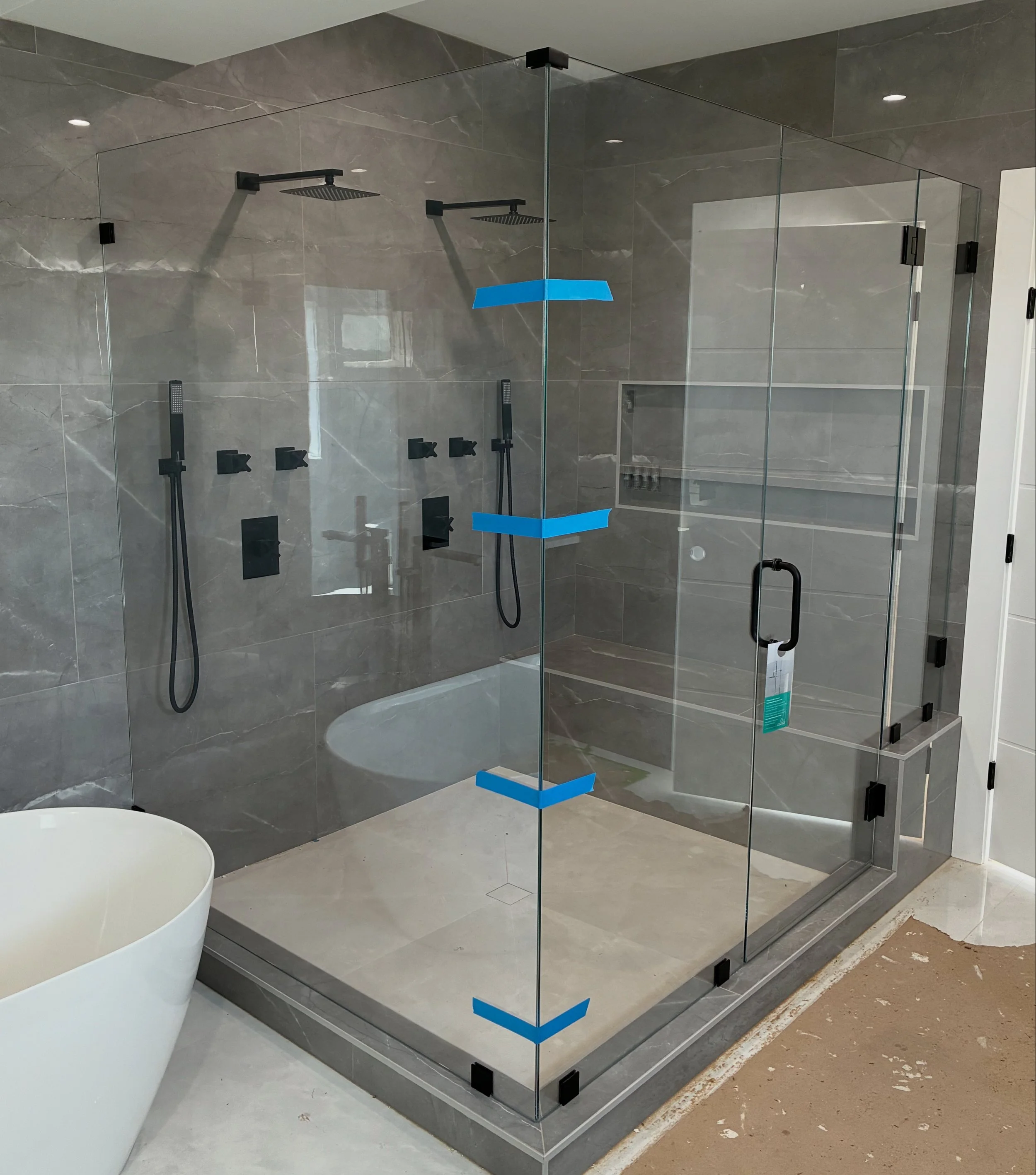 Modern glass shower enclosure with two black showerheads and handheld showers inside, located in a bathroom with gray marble walls, a white bathtub outside, and a door on the right.