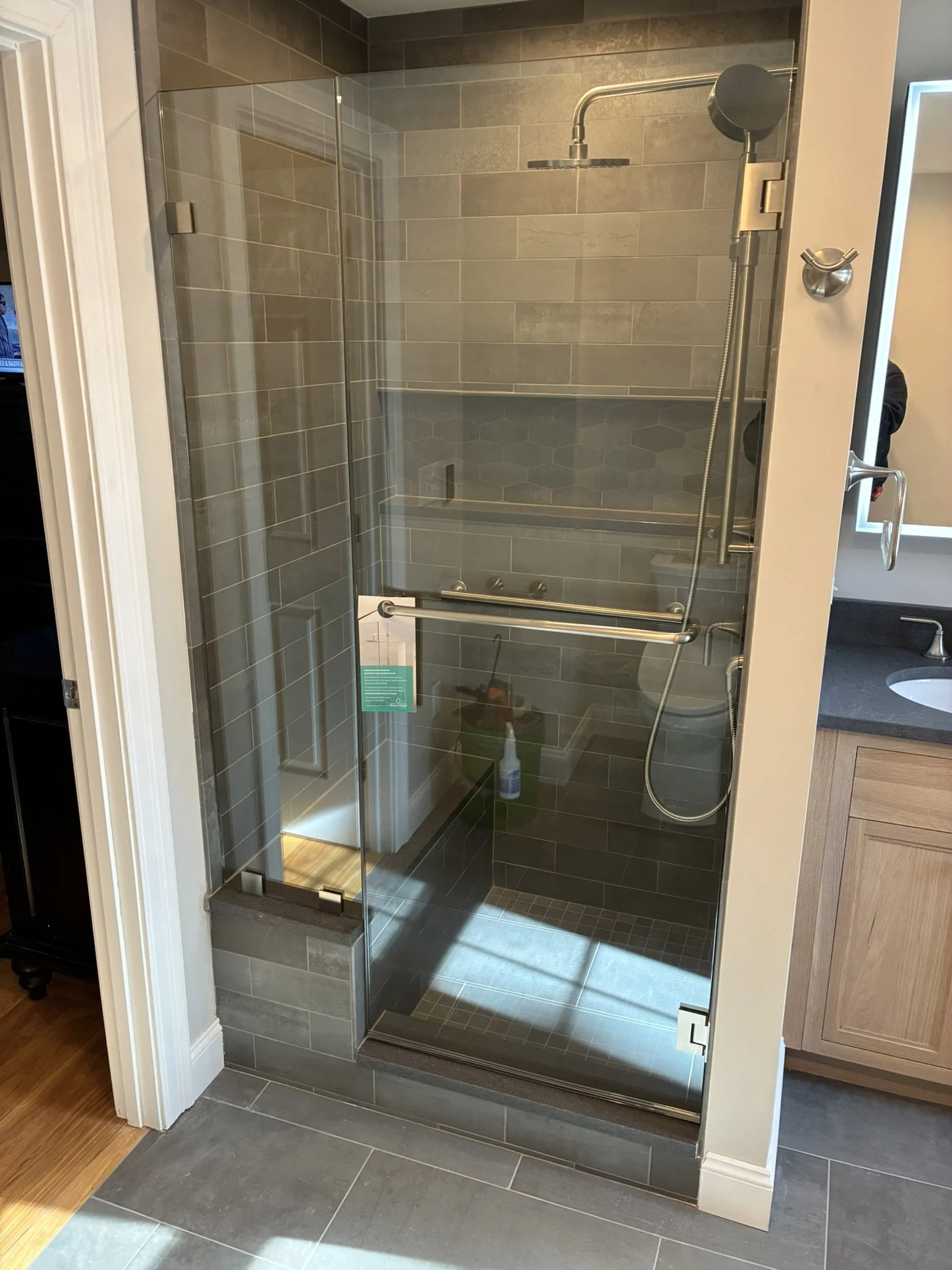 A glass-enclosed walk-in shower with dark gray tile walls and a built-in bench. The shower has a ceiling-mounted rain showerhead and a handheld showerhead attached to a vertical bar. There is a towel bar on the glass door, and the shower area is adja