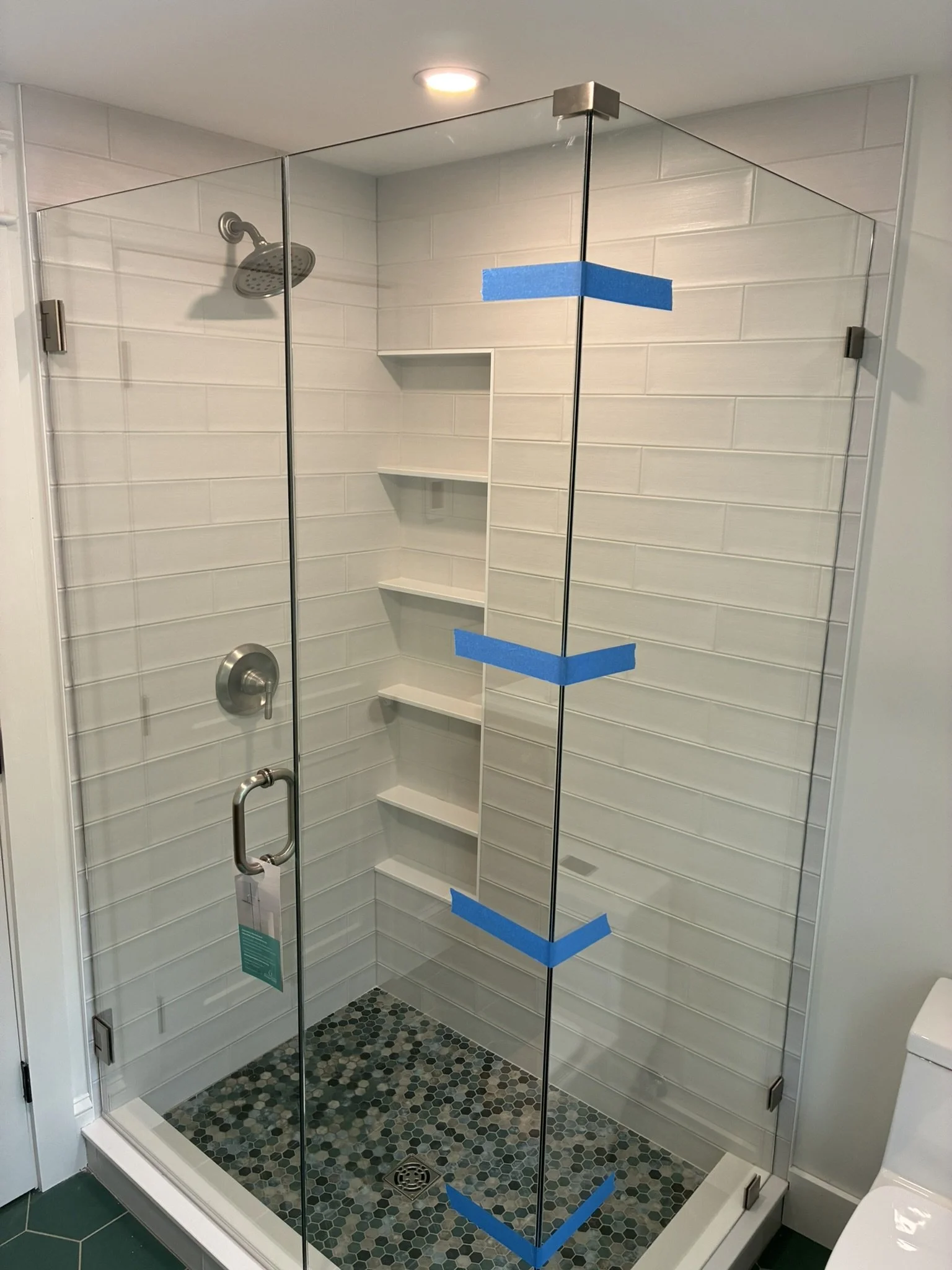 A glass shower enclosure with tiled walls and a built-in corner shelf. The shower has a rain shower head and is marked with blue painter's tape on the door.