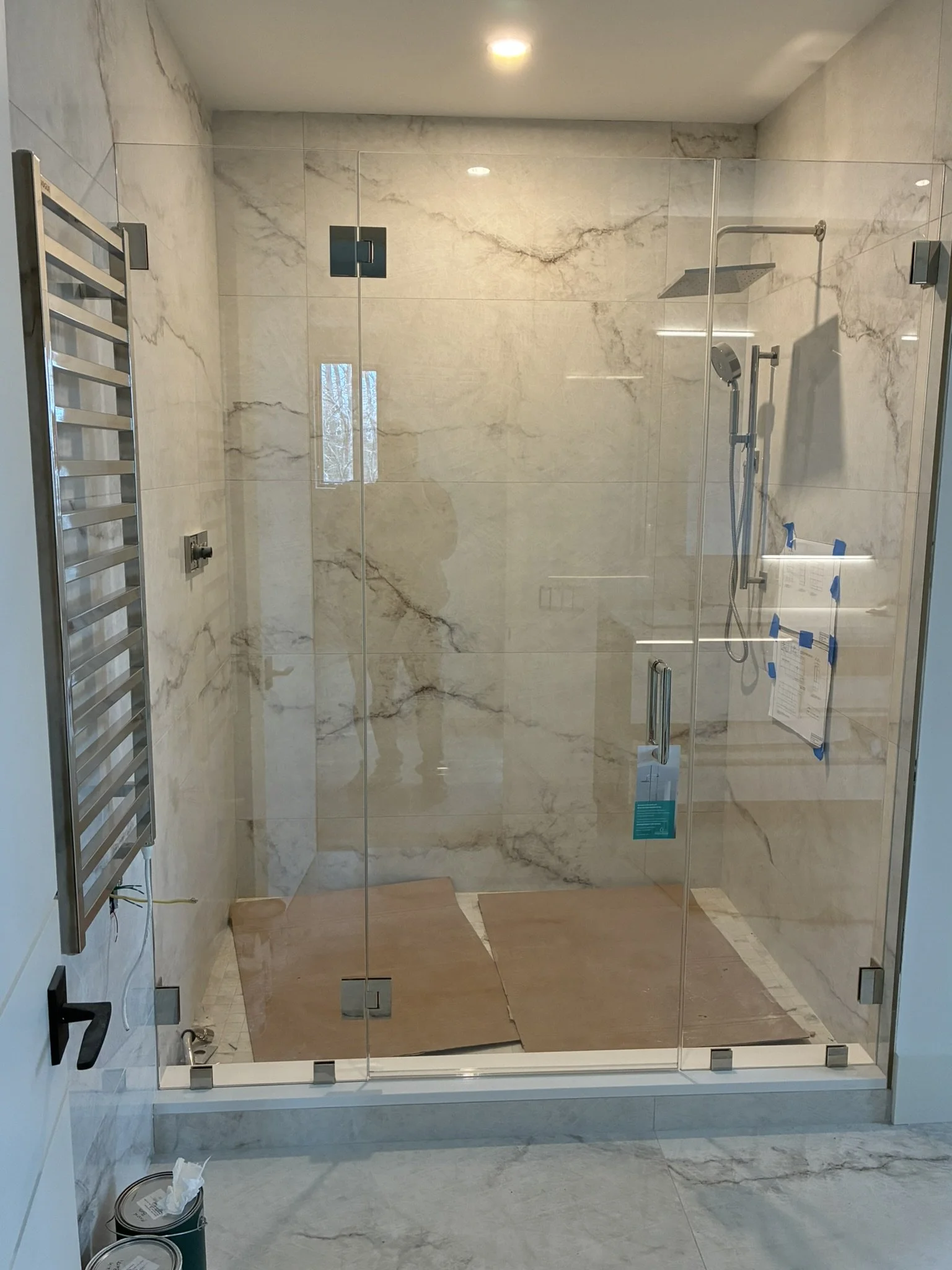 A modern walk-in shower with clear glass doors, marble walls, a rainfall showerhead, a handheld showerhead, and a window on the marble wall. There are some protective blue tape and notes on the right wall. A towel radiator is mounted on the left wall