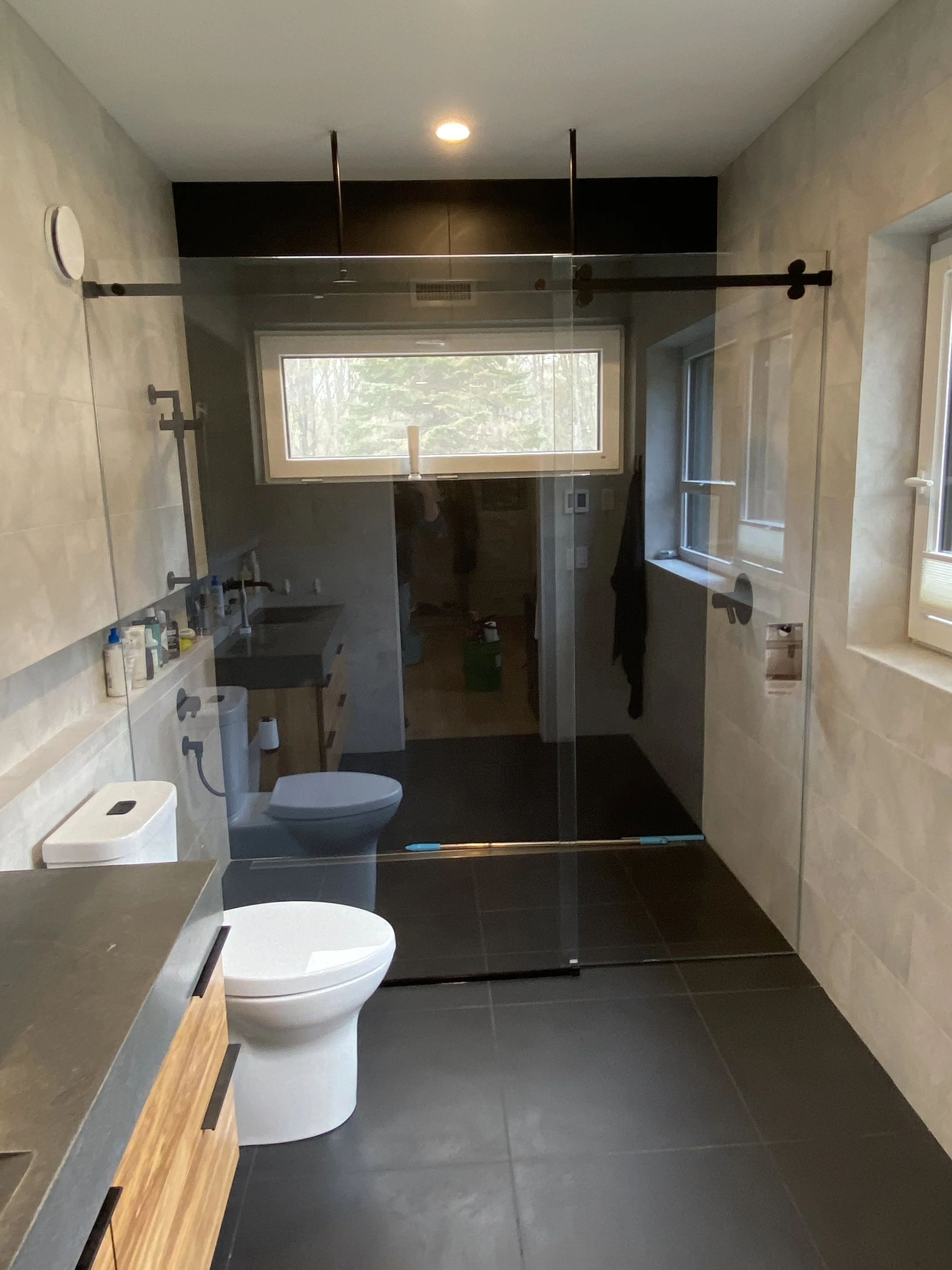 Modern bathroom with glass shower door, toilet, small sink, and window overlooking trees.