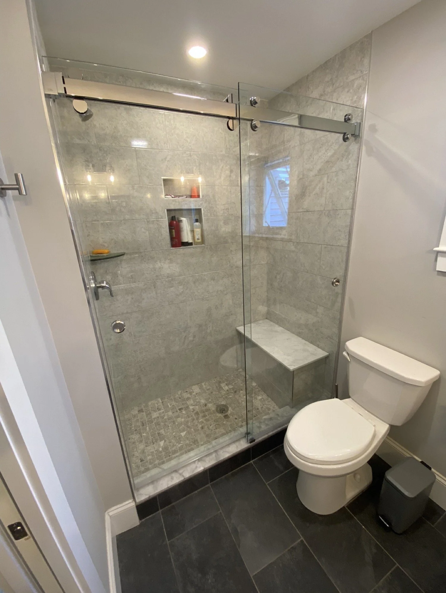 Frameless sliding glass shower door on a chrome roller rail, with marble-look tile, a corner bench, a niche and a window, in an Ogunquit and York area Maine bathroom