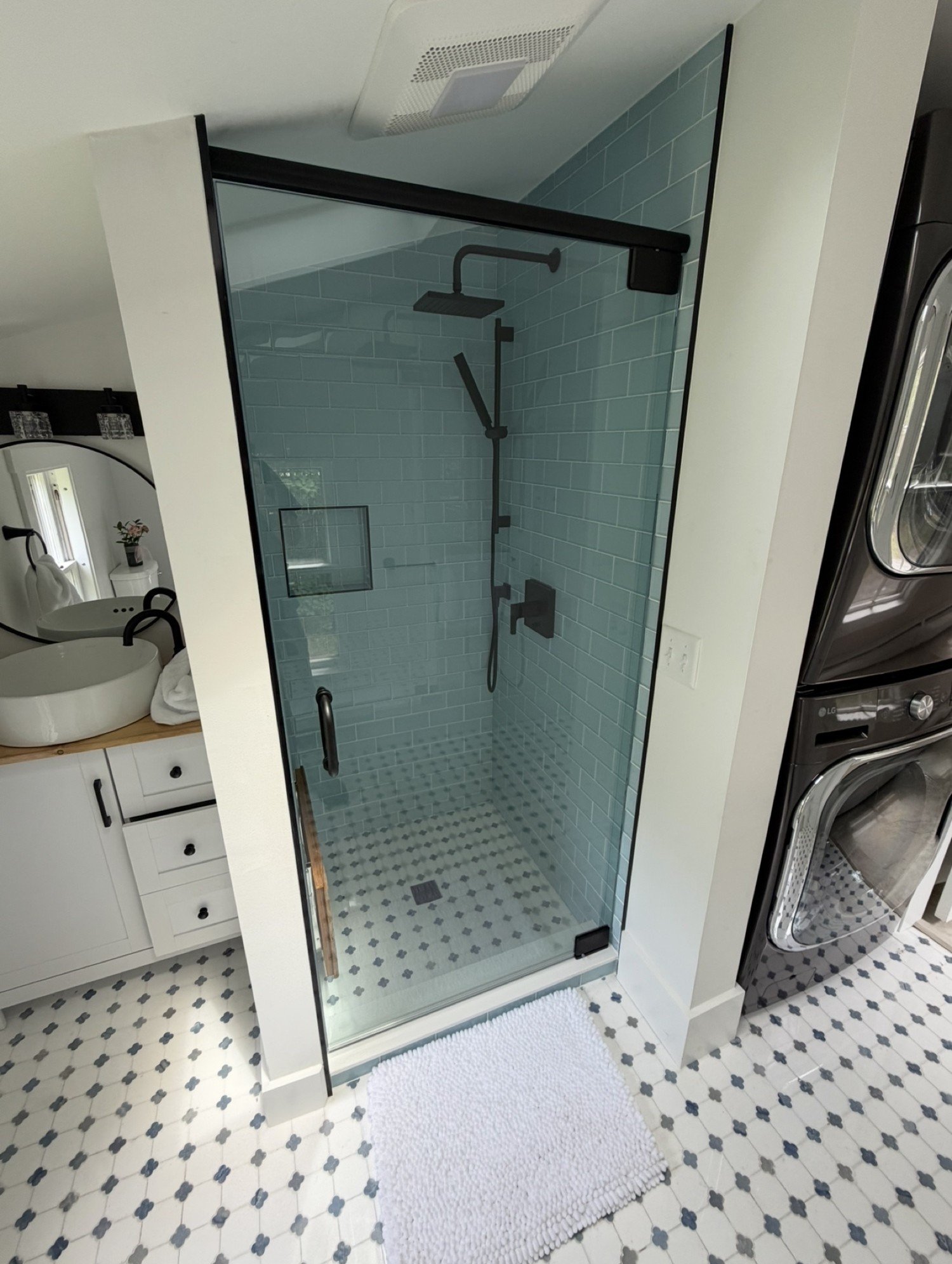 Frameless glass shower door with a matte black header bar, cut to a sloped ceiling, with aqua subway tile, black fixtures and a black and white mosaic floor, in an Ogunquit and York area Maine bathroom