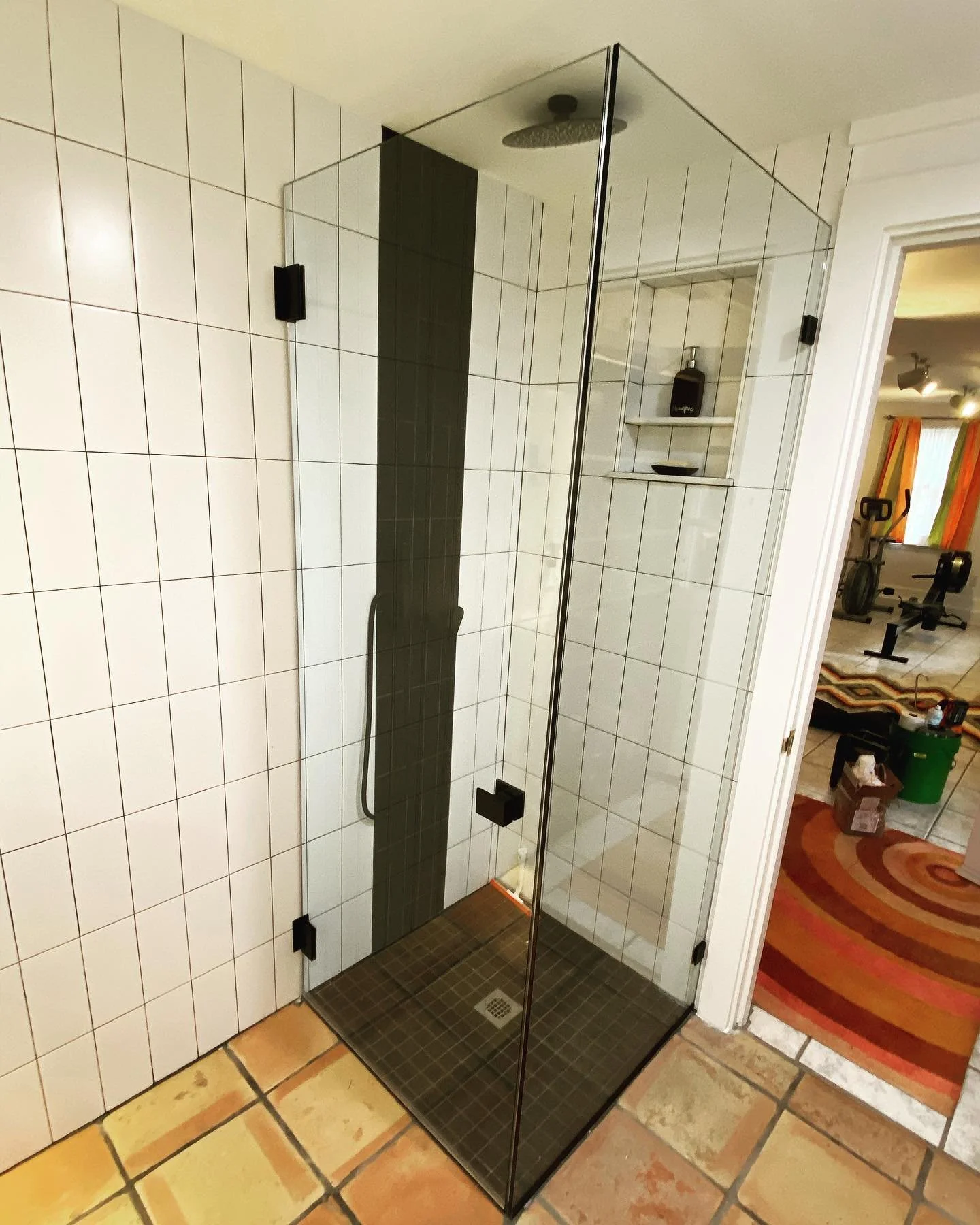 Frameless glass corner shower with a hinged door and panel, matte black hardware, white tile and a black accent stripe in a small Cumberland Foreside, Maine bathroom with terracotta floor tile