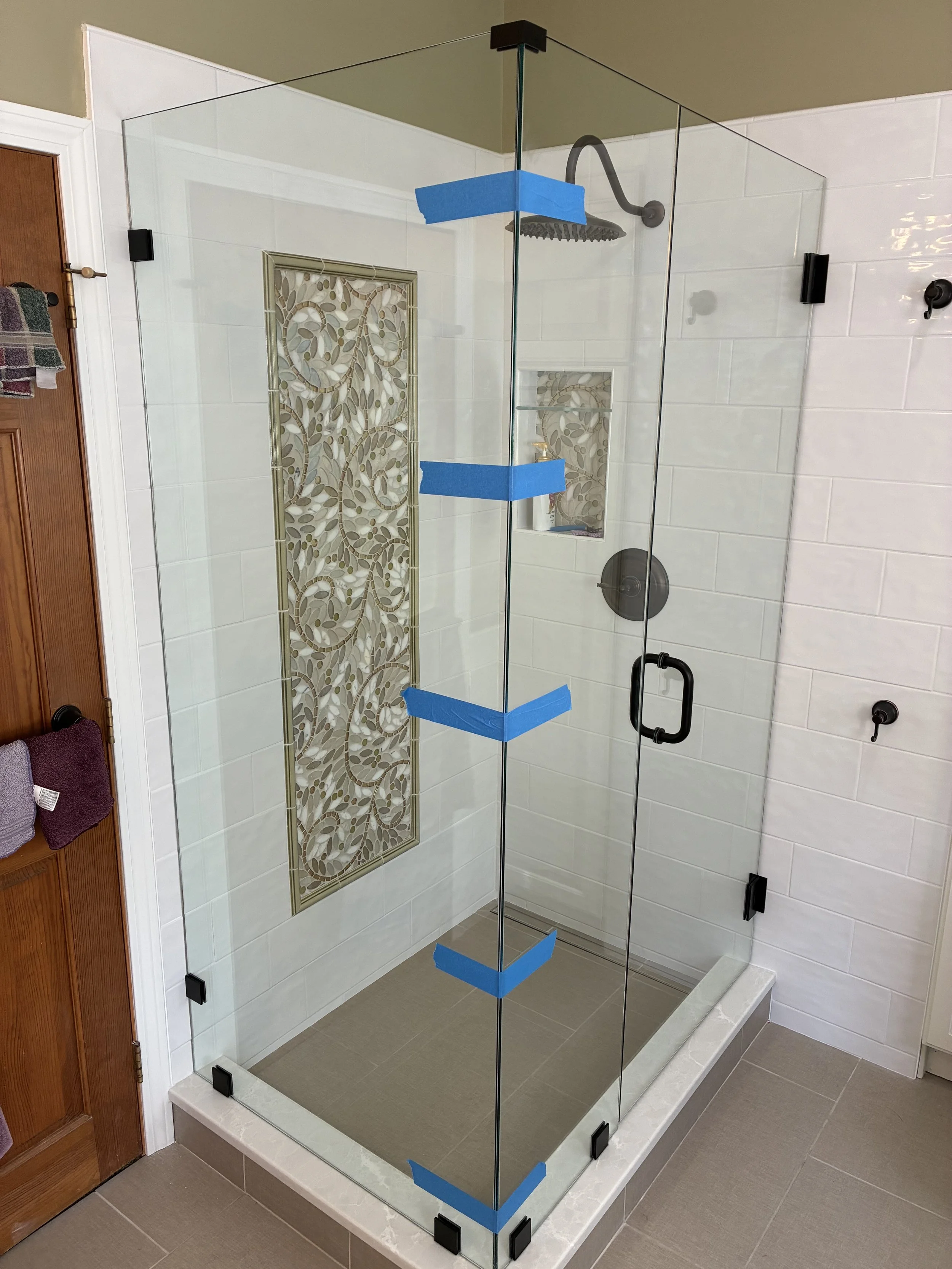 A glass shower enclosure with black hardware and blue painter's tape on the edges, inside a bathroom with beige tiled flooring and white tiled walls, featuring a built-in decorative tile niche with leaf-patterned tile.