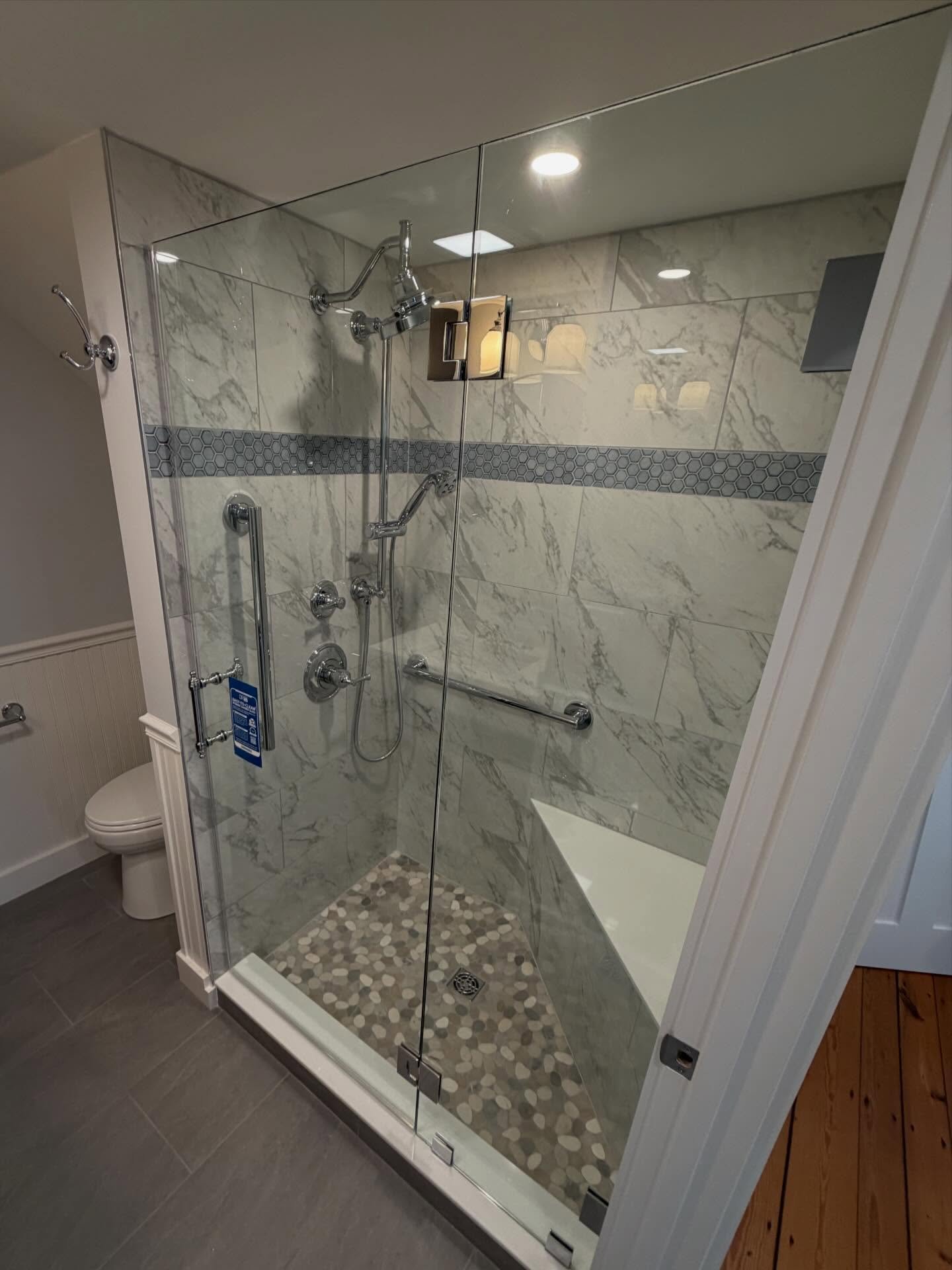 Frameless glass corner shower with a hinged door, inline panel and polished chrome hardware, marble-look tile with a hex mosaic band, pebble floor and bench in a Biddeford Pool, Maine home
