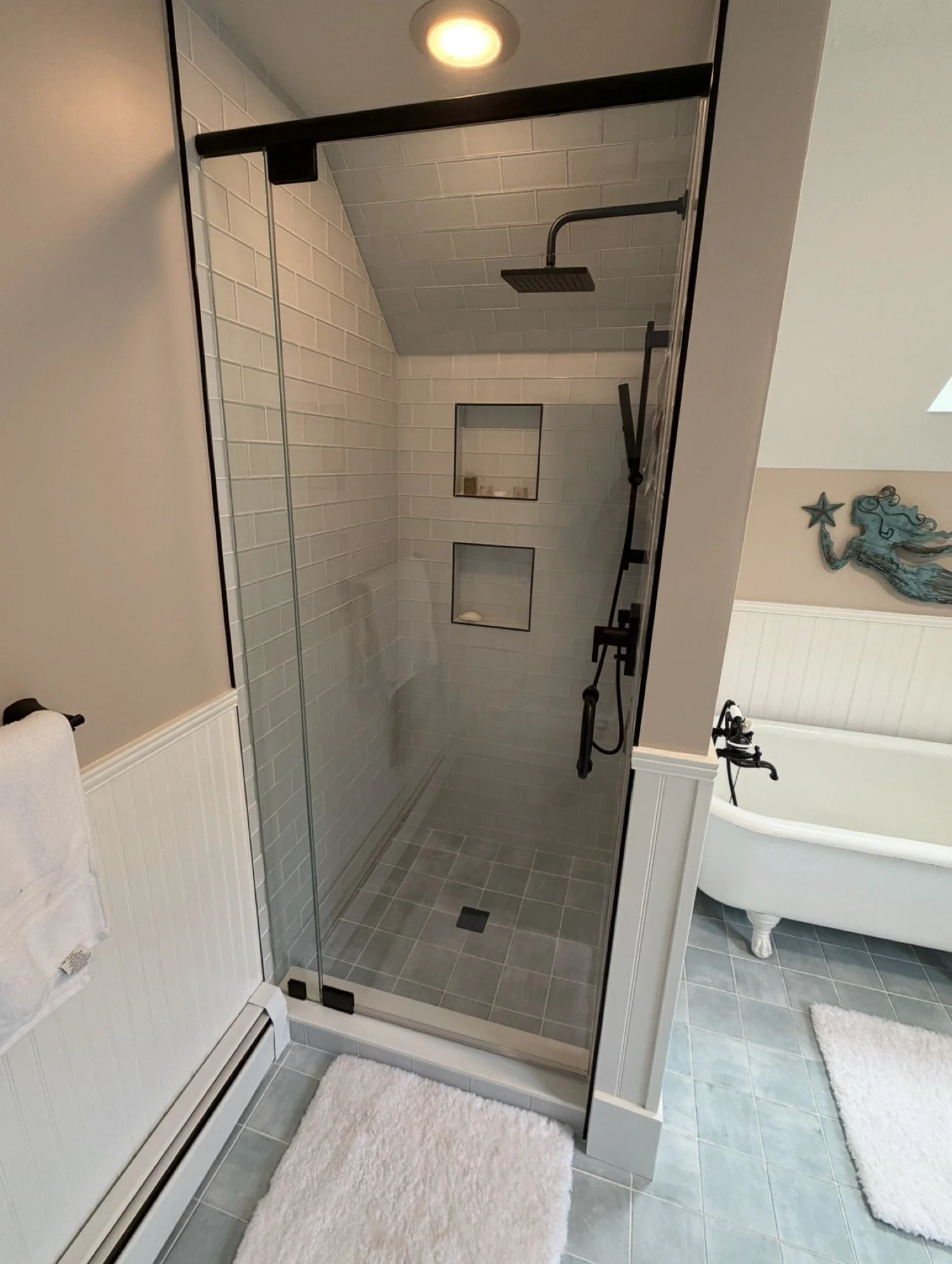 Frameless glass shower door and panel with a matte black header under a sloped ceiling, white subway tile and two niches, beside a clawfoot tub, in an Ogunquit and York area Maine bathroom