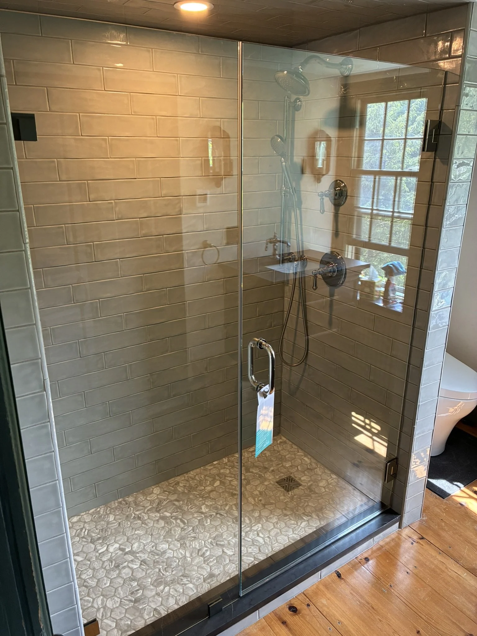 A walk-in shower with glass doors, beige subway tile walls, a rainfall showerhead, and a handheld showerhead. The floor has pebble tiles, and a large window with a view of trees is reflected in the glass. A toilet is partially visible outside the sho