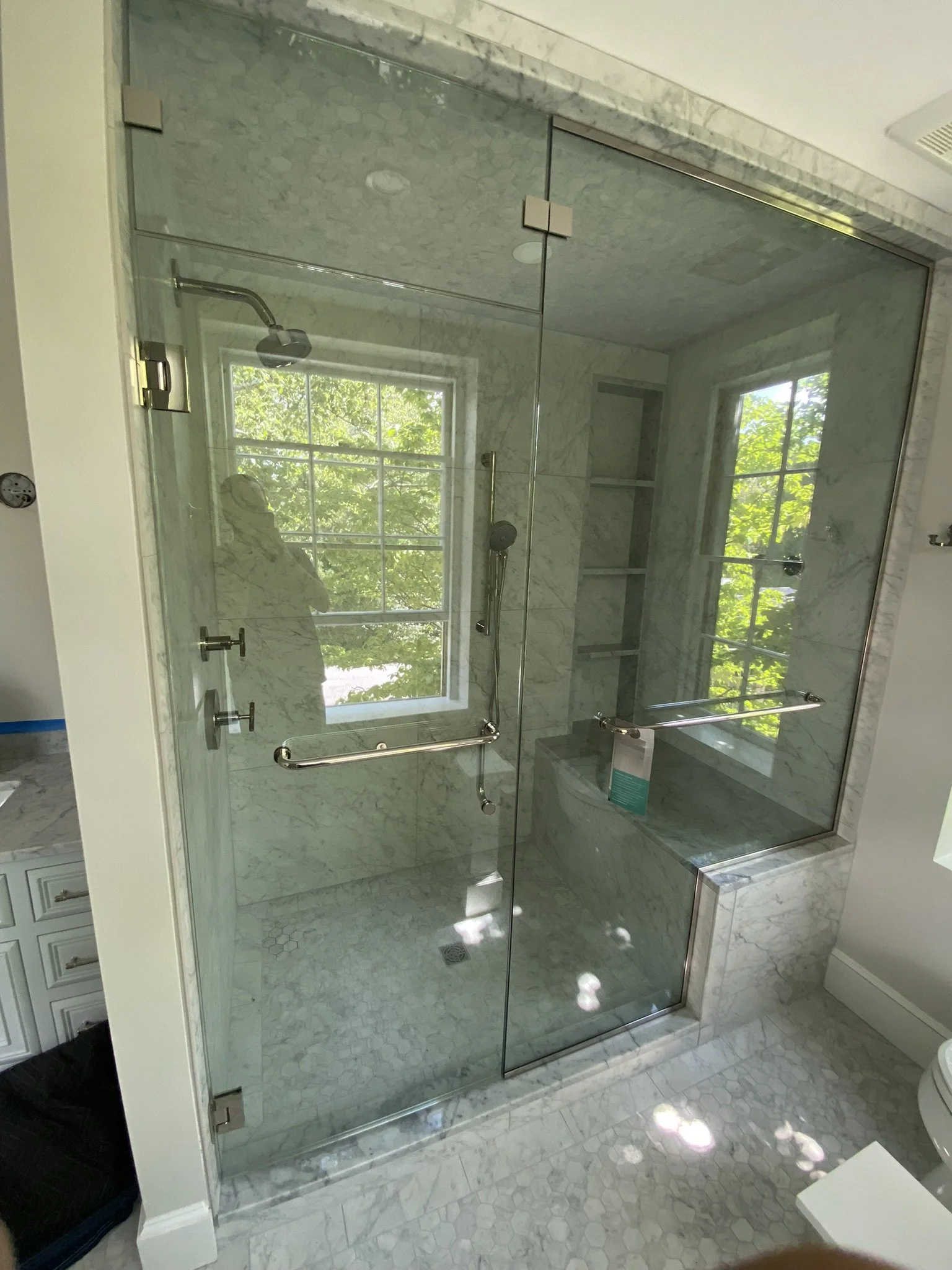Glass shower enclosure with marble tiles, built-in marble bench, two windows, and chrome fixtures, in a bathroom.