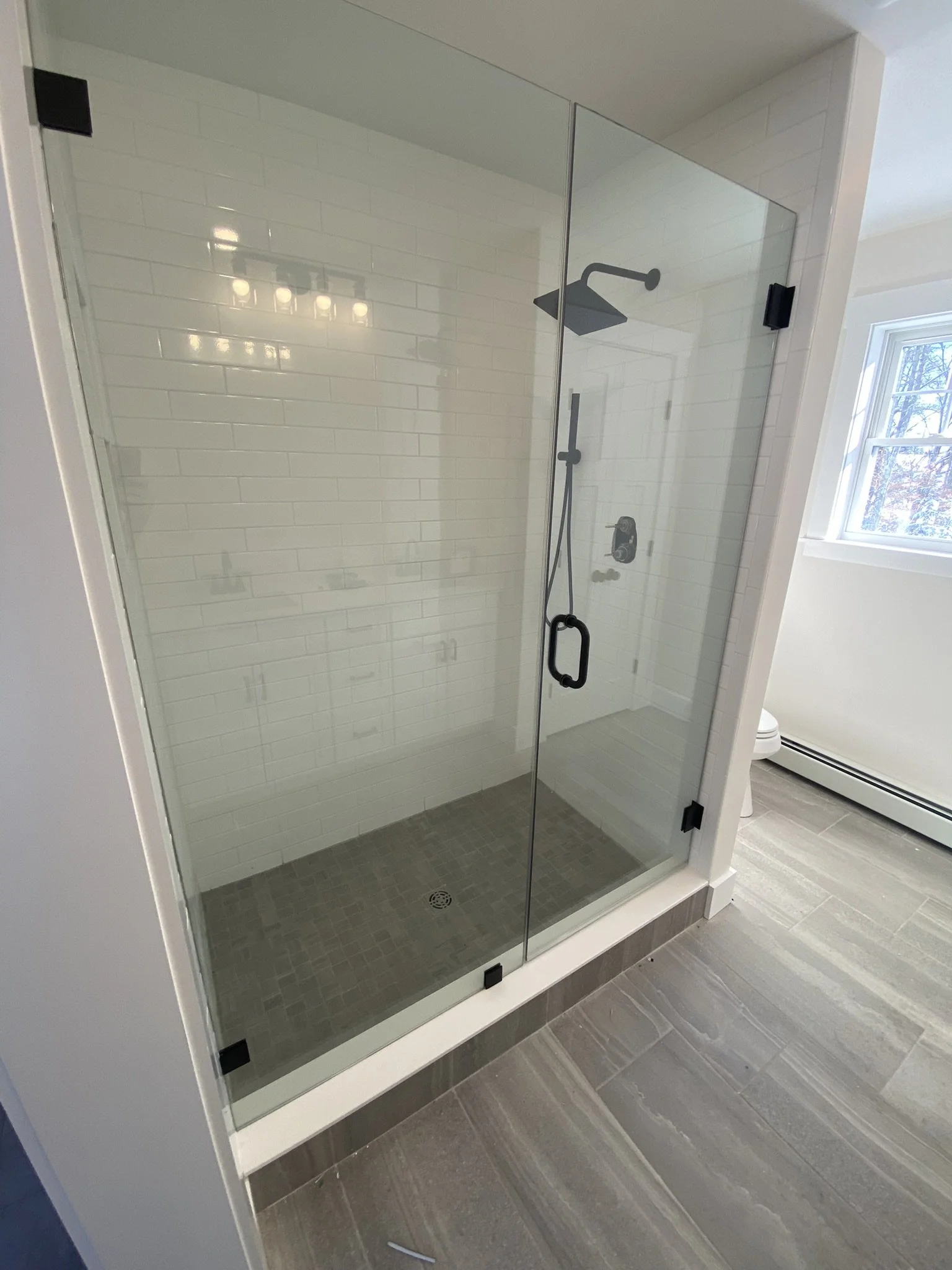 Glass shower enclosure with a black handle and a showerhead, white tiled walls, and a gray tiled floor.