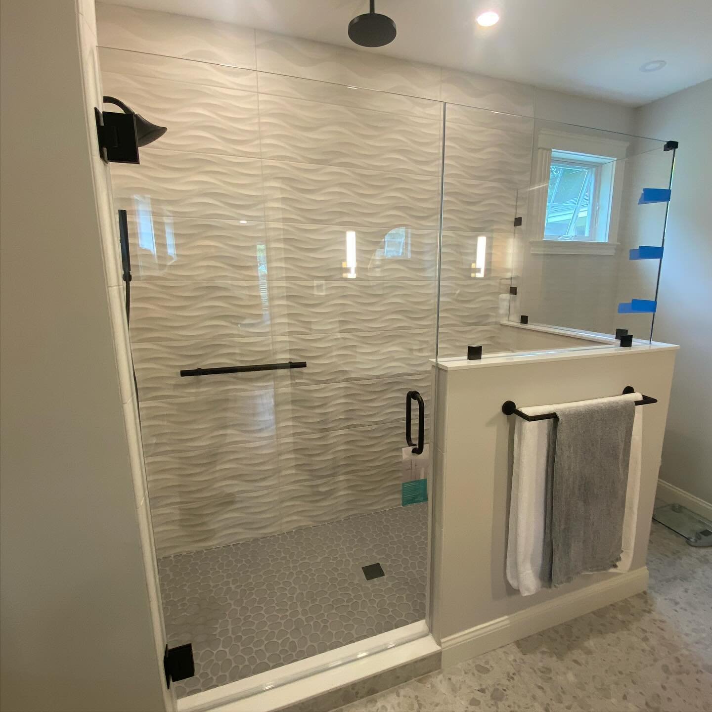 Frameless low-iron glass shower enclosure with a hinged door, inline panel and a panel on a half wall, matte black hardware and a towel bar, against white wave-pattern tile in a Falmouth, Maine bathroom
